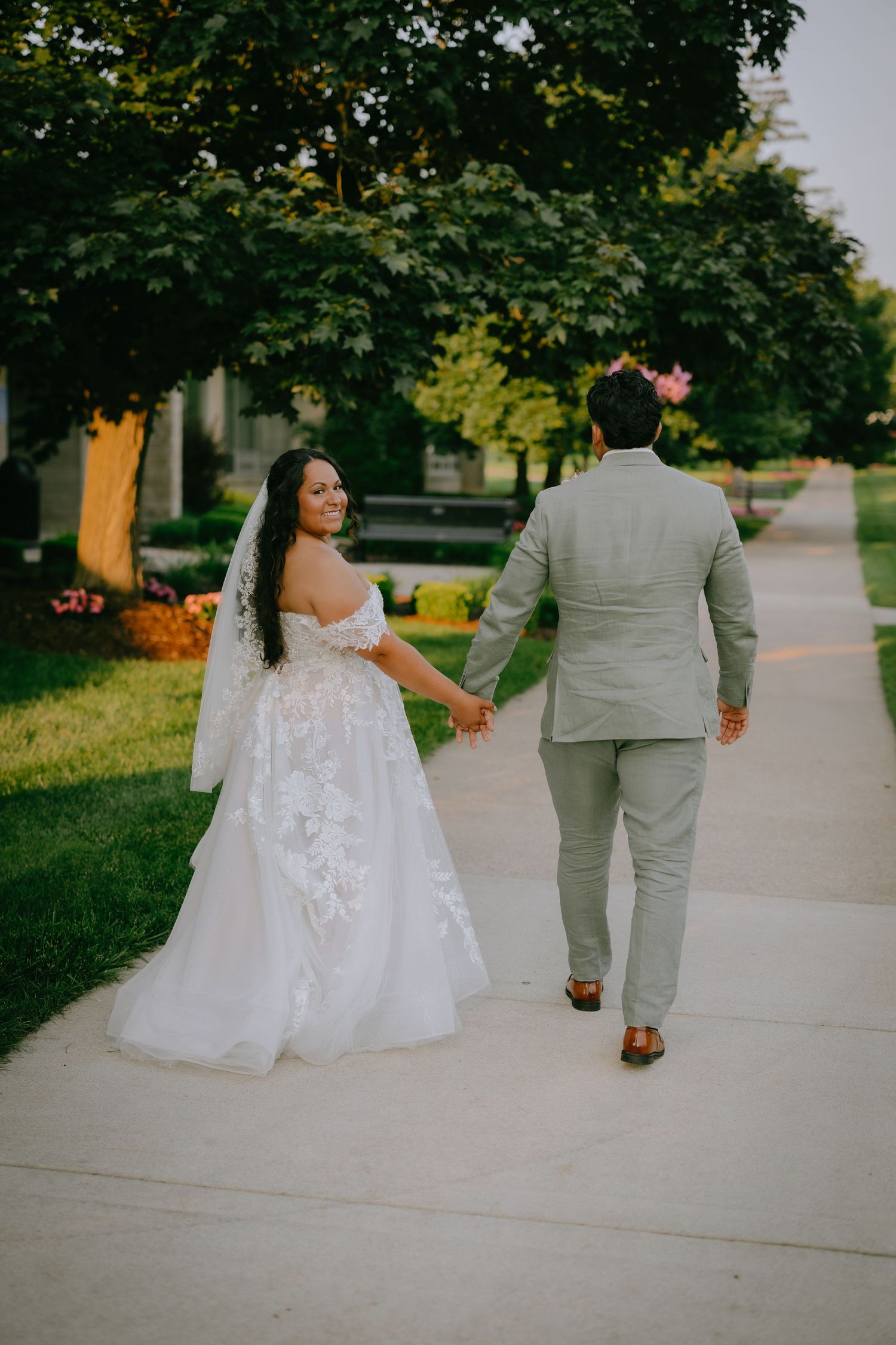A wedding couple walks along a tree-lined sidewalk on their special day, with the bride in a white lace gown.