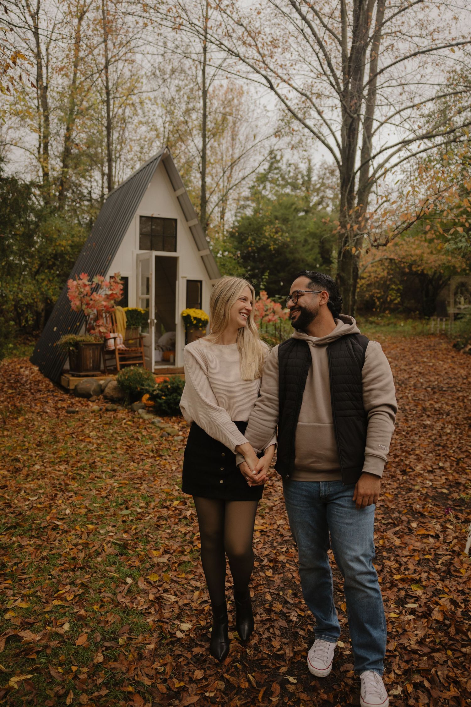 A couple walking through fallen autumn leaves near a small wooden structure in the woods.