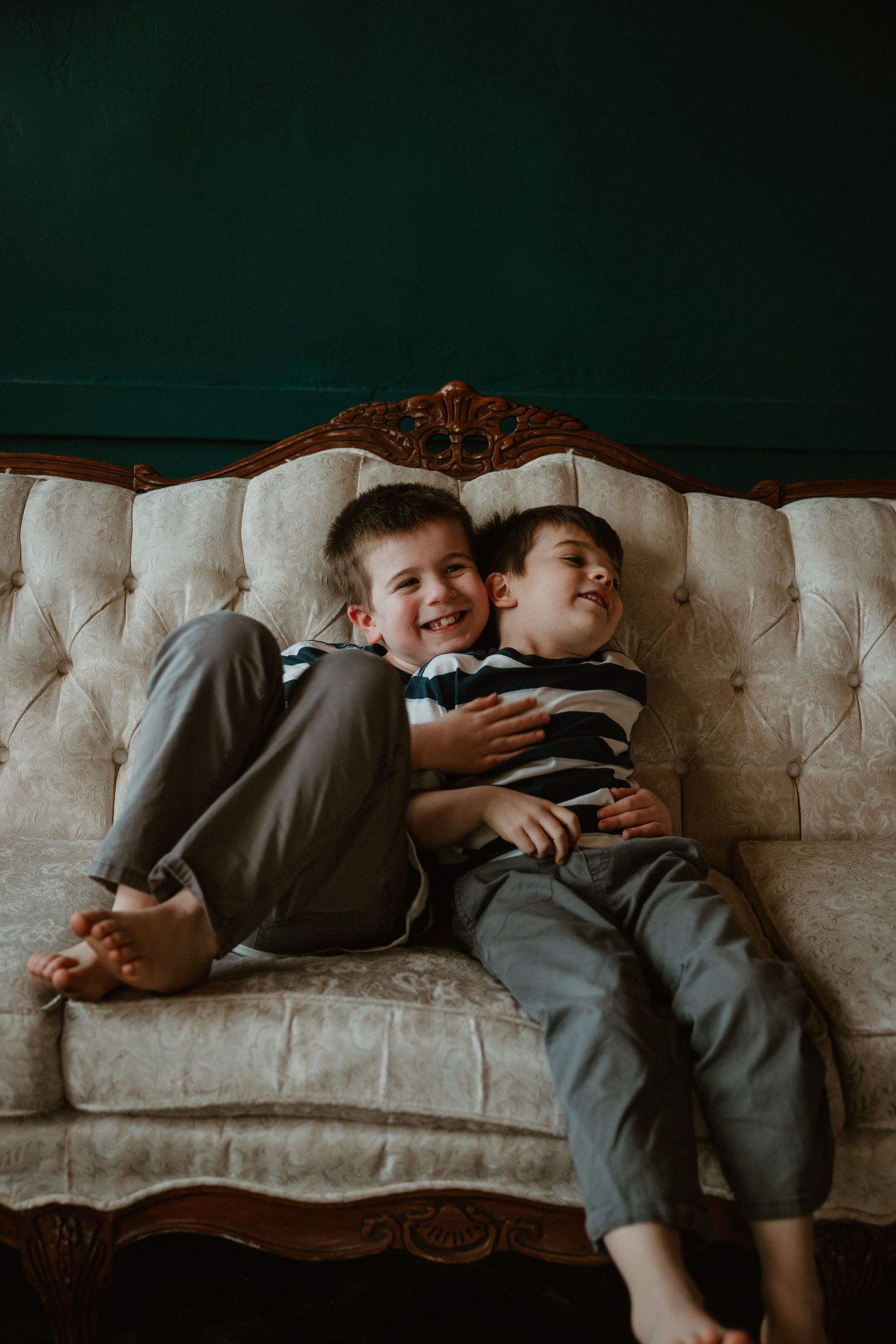 Two young boys laughing together on an elegant tufted vintage couch against a dark green wall.