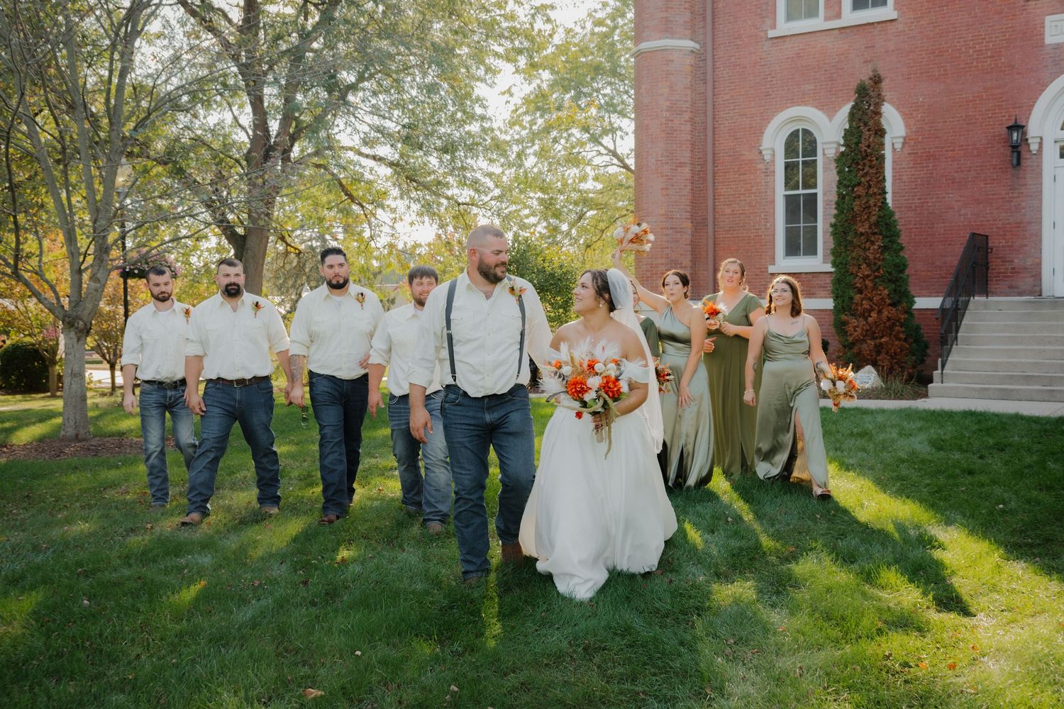 Wedding party walks outdoors near brick building, bride in white gown with bouquet, groomsmen in jeans, bridesmaids in sage green.
