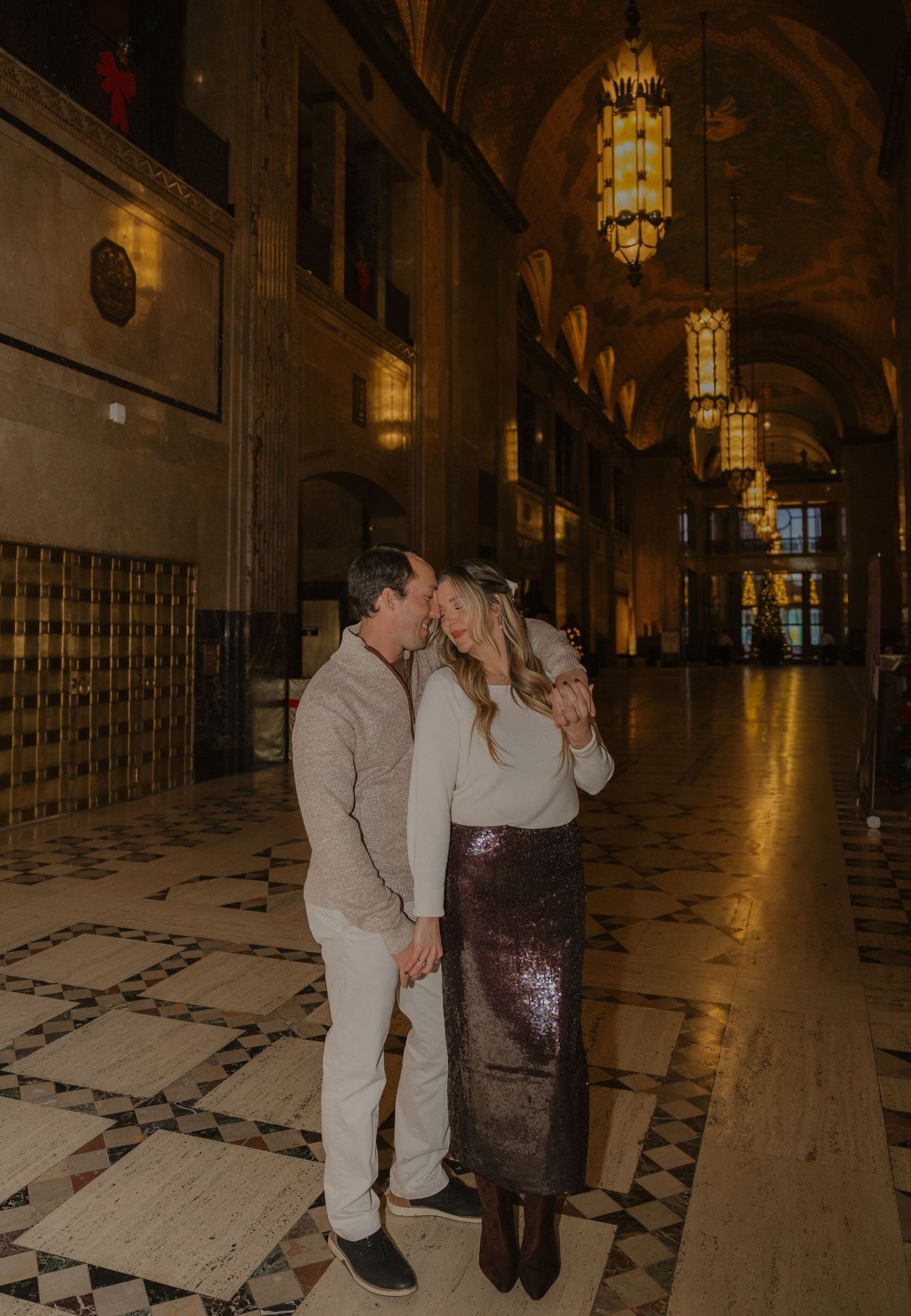 Couple sharing a romantic moment in an elegant grand hall with ornate chandeliers and marble floors.