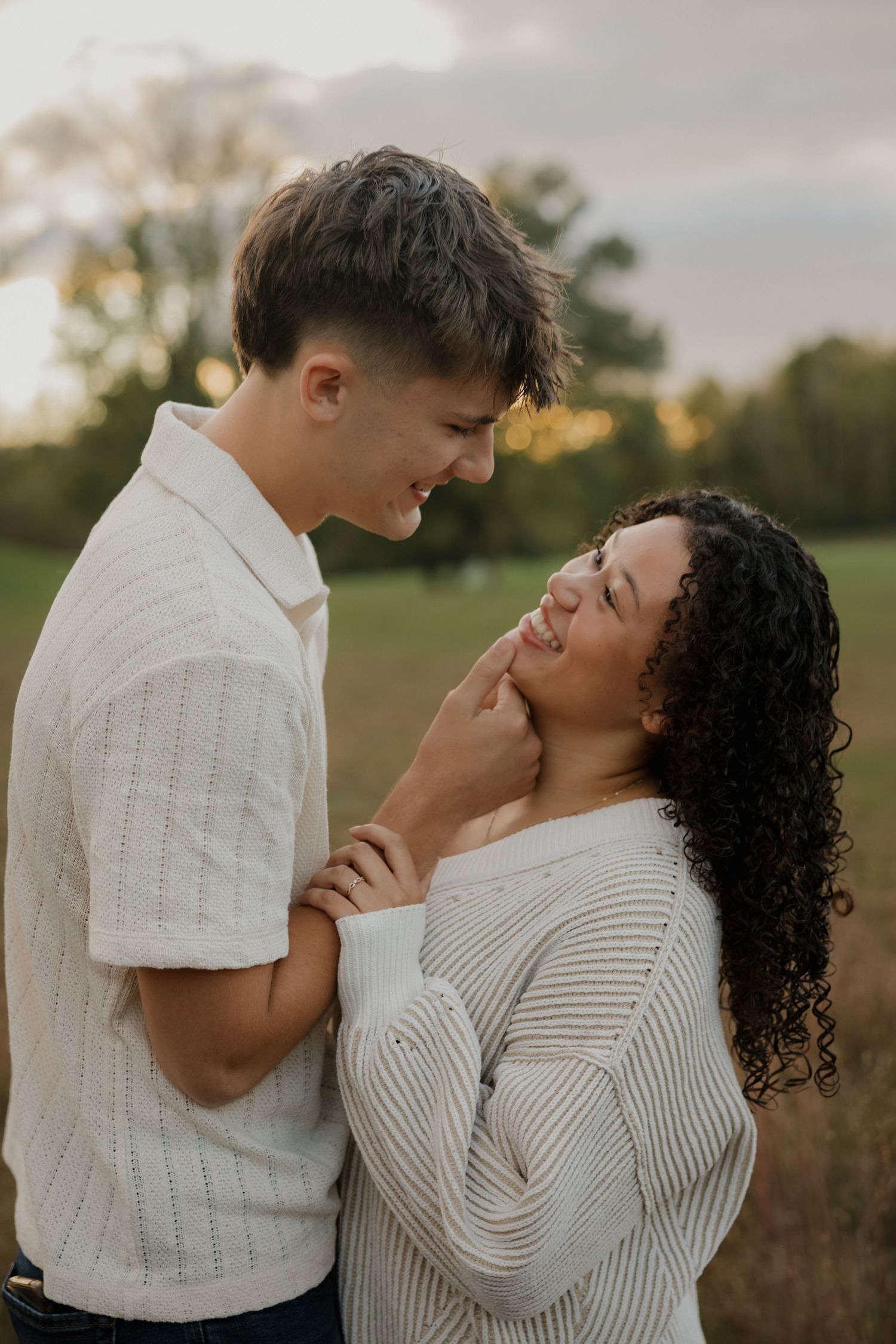 A tender moment between a couple wearing white sweaters sharing a laugh in a sunset field.
