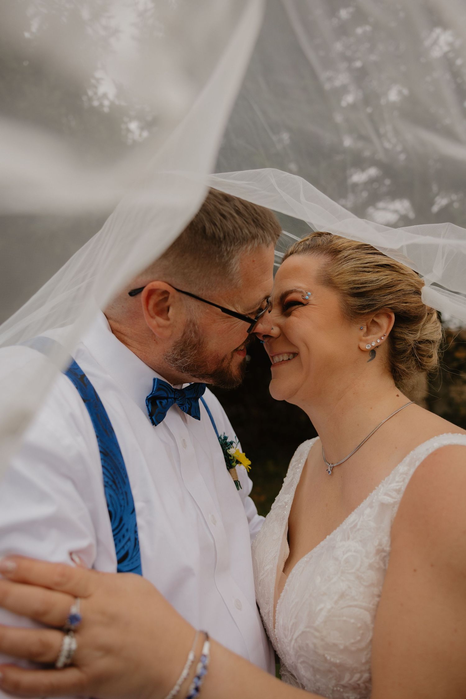 Bride and groom share a romantic moment under a flowing veil, smiling at each other in wedding attire.