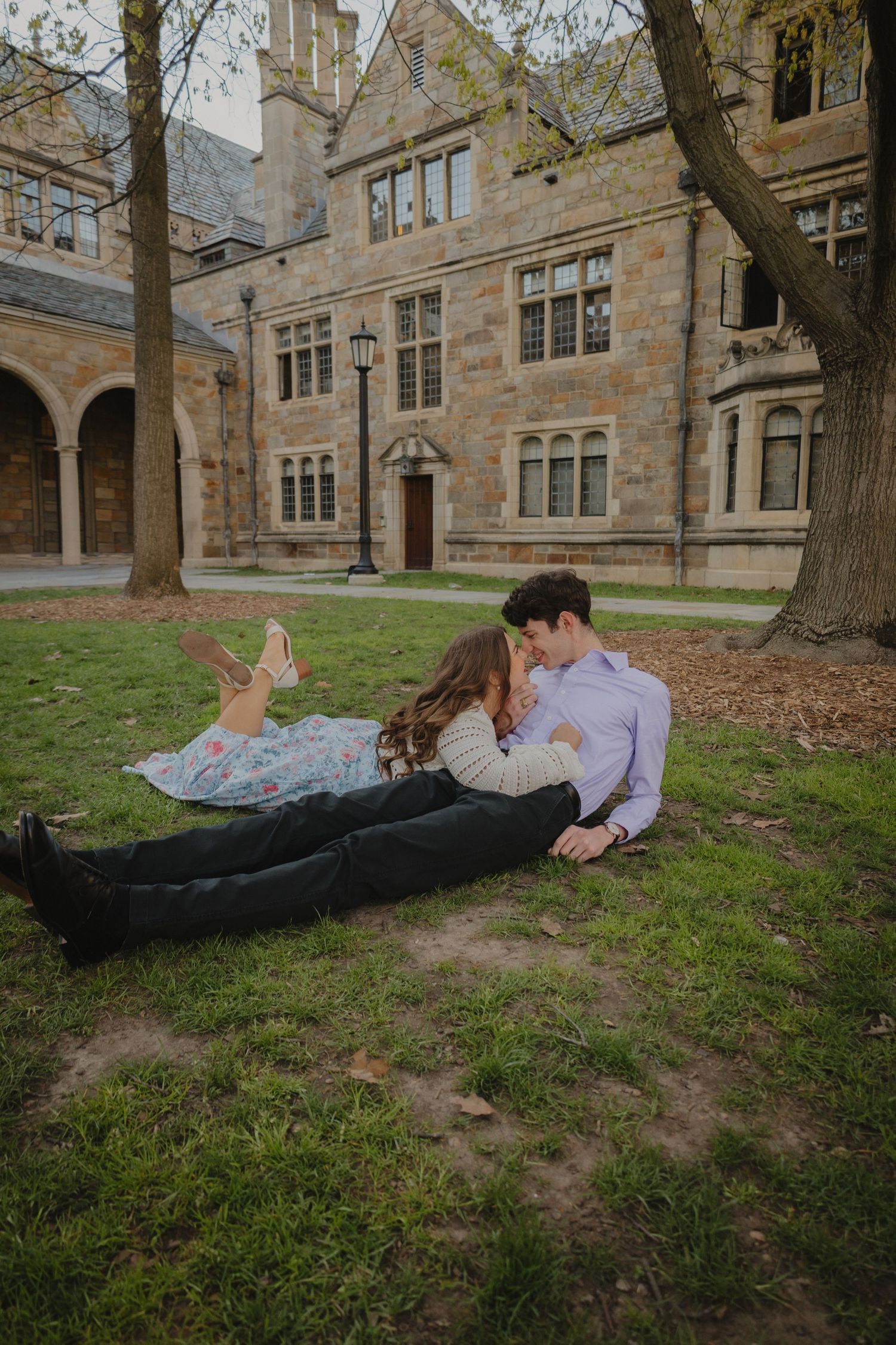 Couple laying on grass in romantic embrace outside historic stone university building during engagement photo session.