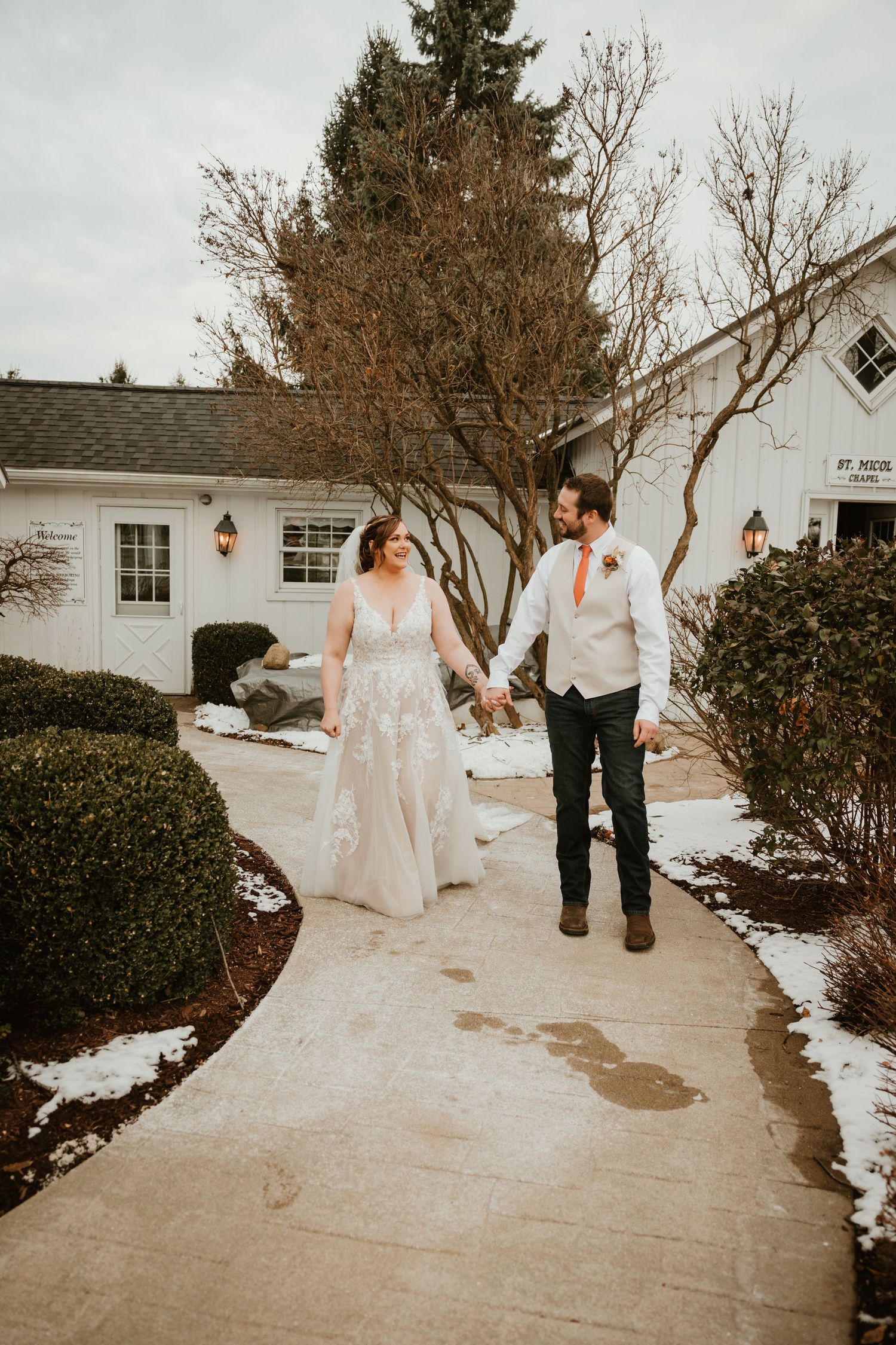 A winter wedding portrait showing a couple walking on a snowy path near a white building.