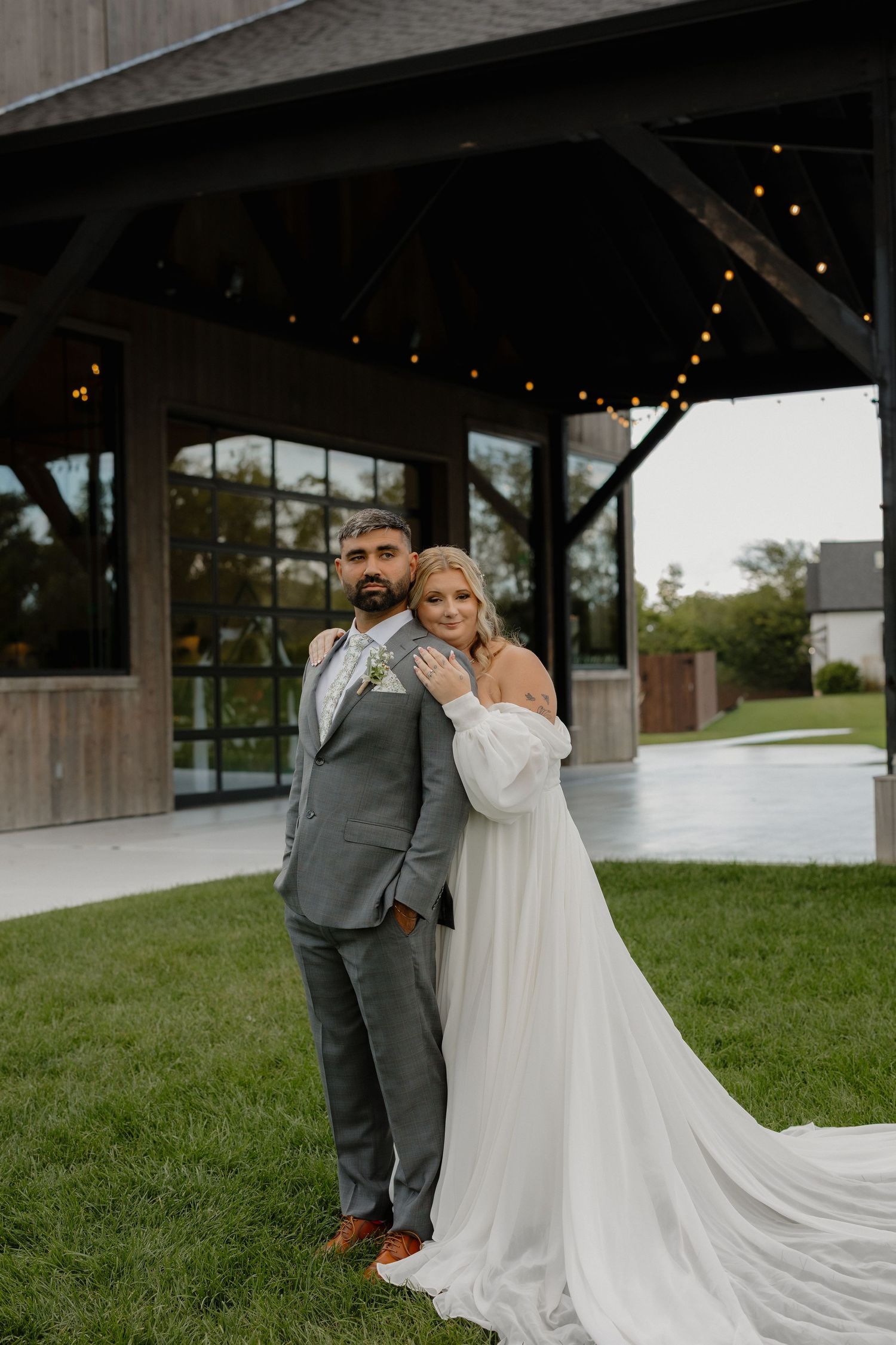 Wedding couple in white gown and gray suit embracing outside rustic pavilion venue.