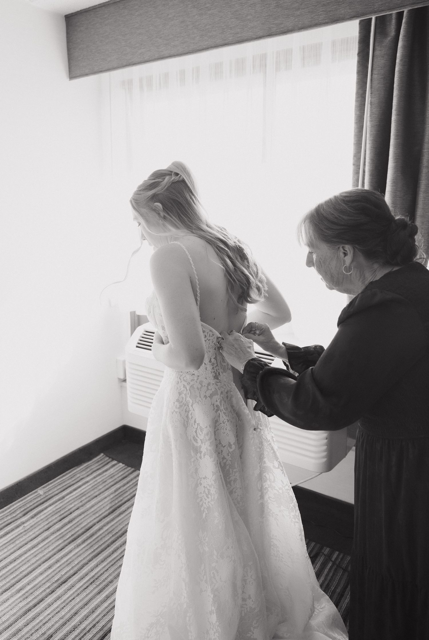 Black and white photo of a bride in lace wedding gown being helped with her dress by a woman in a hotel room.