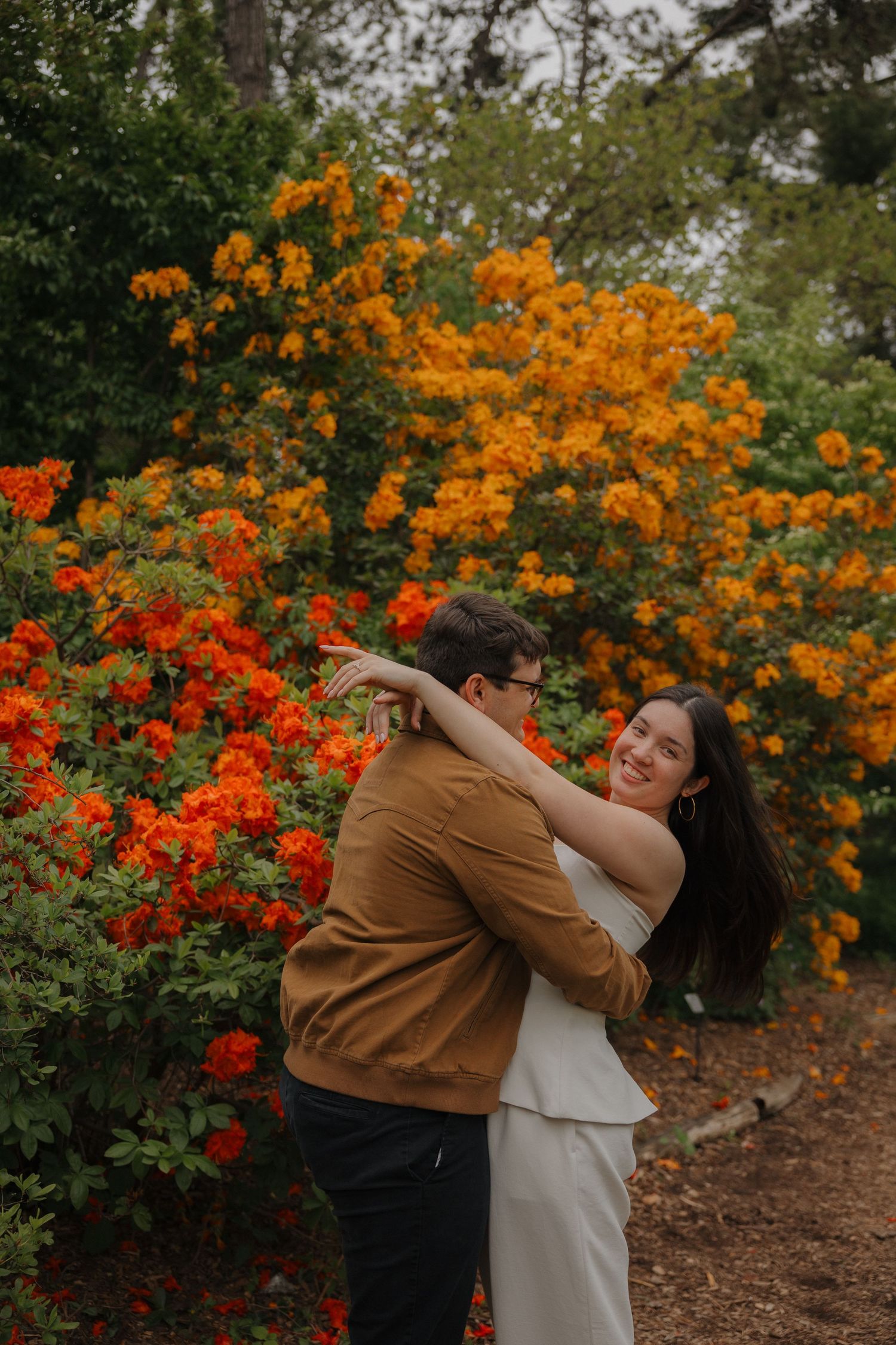 Couple embracing and smiling in front of vibrant orange and red flowering bushes in a lush garden setting.