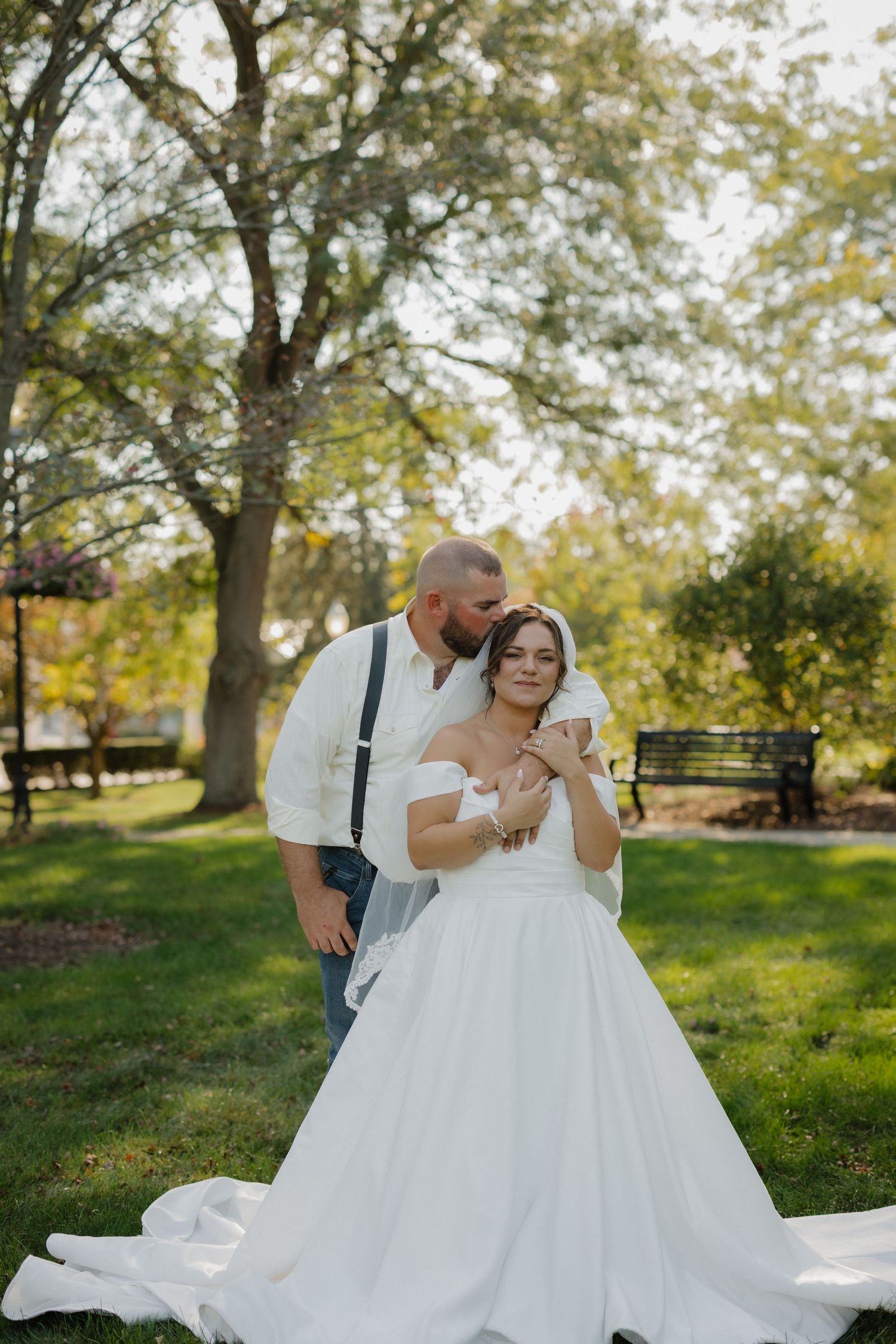 Bride in white off-shoulder gown with groom in suspenders embracing outdoors in a lush green park setting.