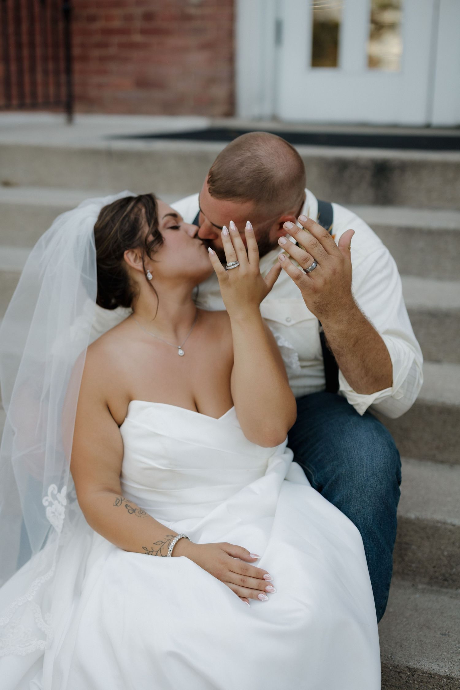Bride and groom share a romantic kiss on steps, bride in strapless white gown with veil, groom in white shirt and jeans.