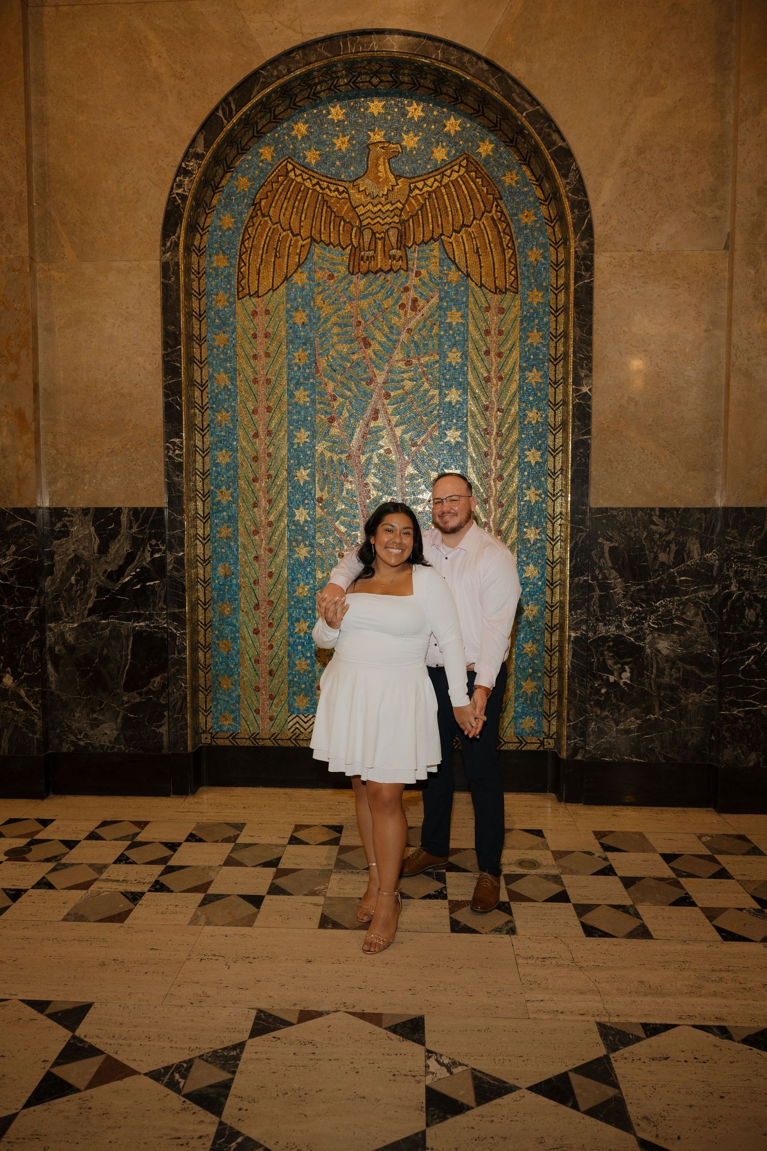 Couple posing in front of ornate mosaic mural with eagle motif in elegant marble-floored building interior.