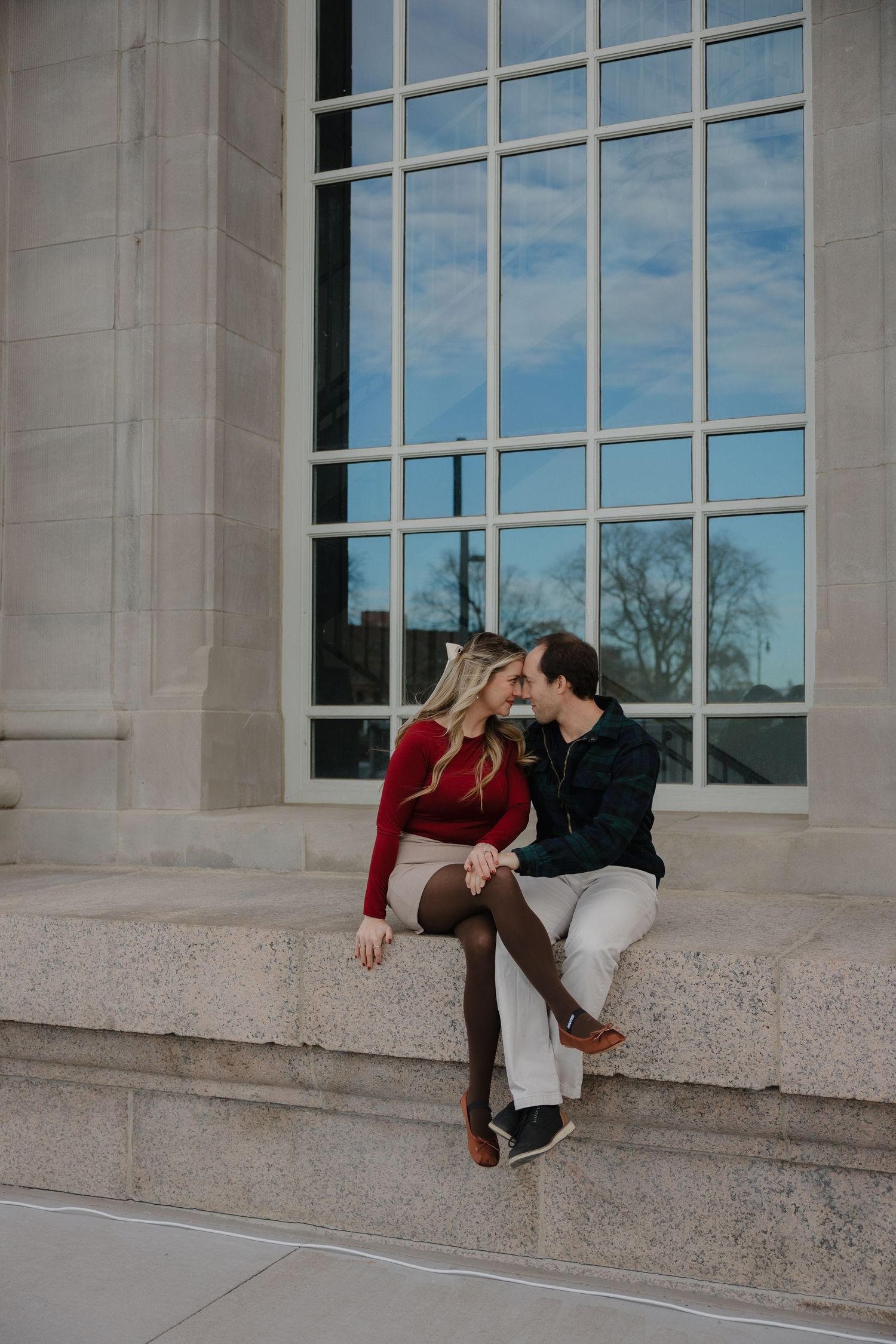 Couple sharing a romantic moment sitting on stone steps outside a large building with tall windows on a cloudy day.