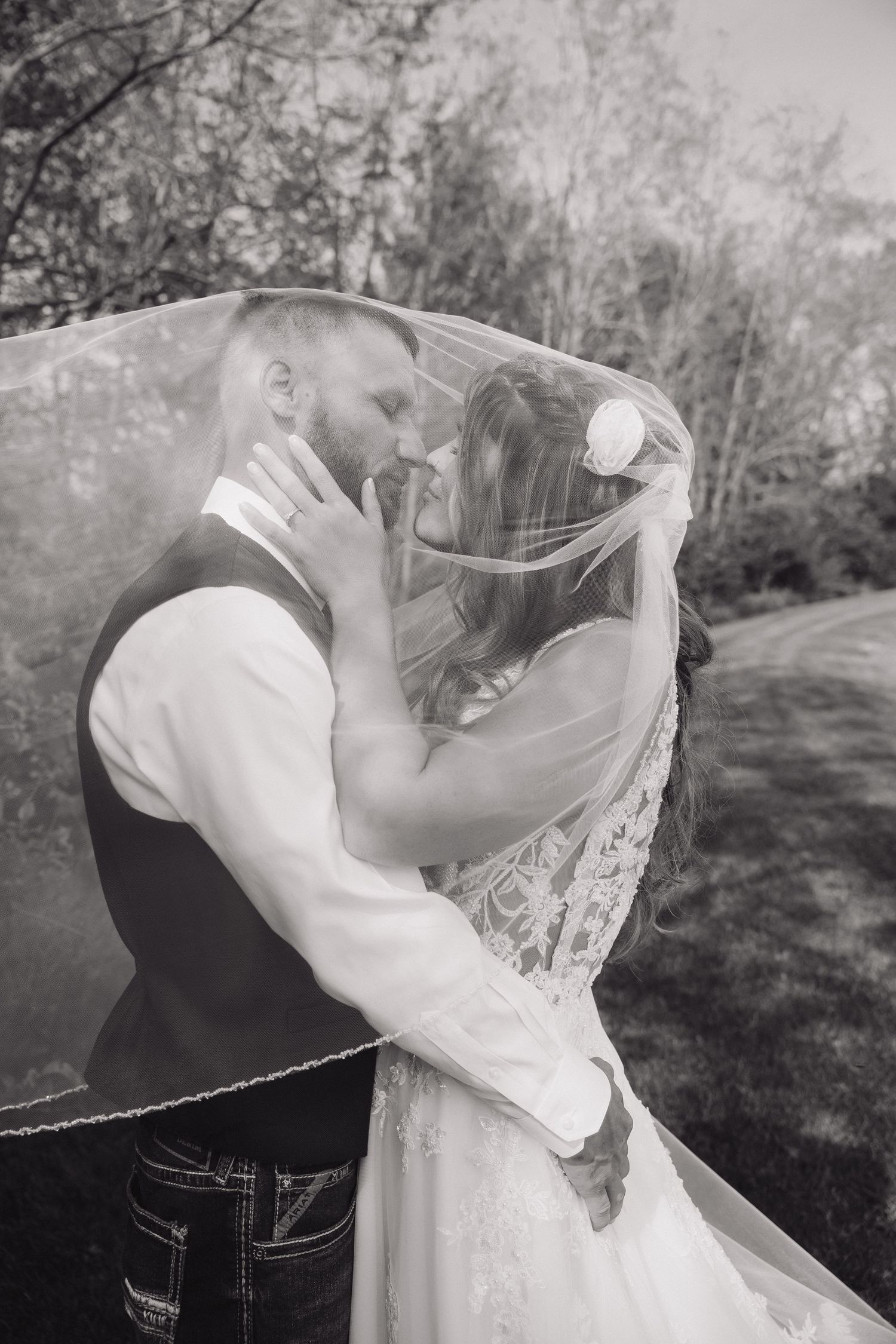 Black and white photo of bride and groom sharing romantic moment under wedding veil outdoors.