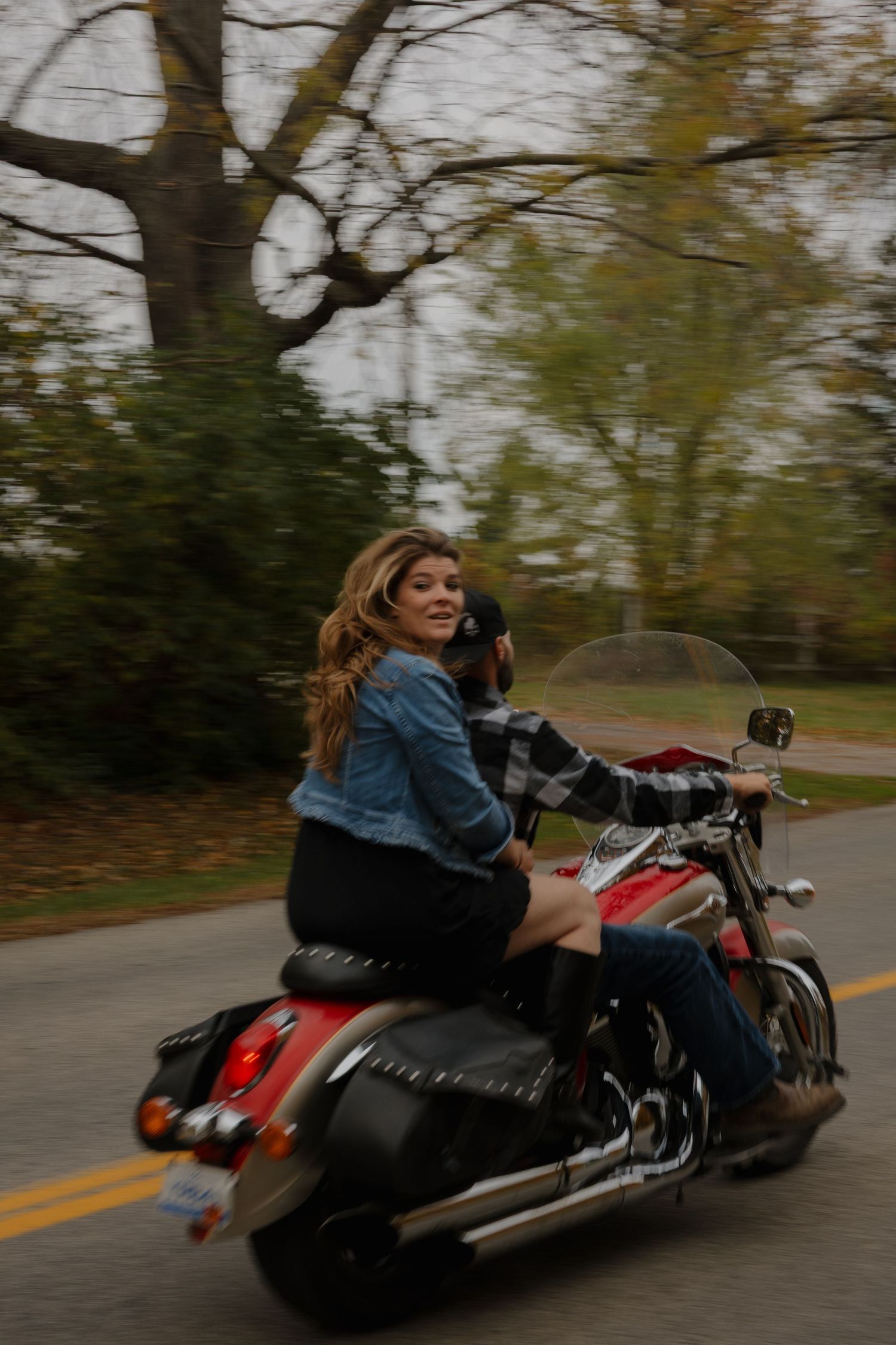 A motorcyclist in denim jacket rides a black and red cruiser motorcycle on a tree-lined autumn road.