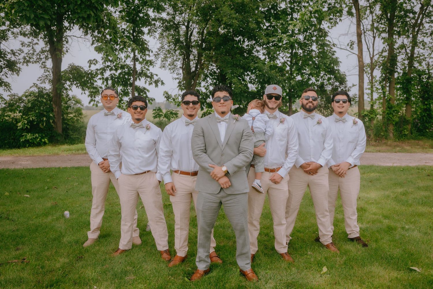 A groomsmen party poses in coordinated light-colored attire on a green lawn with trees in the background.