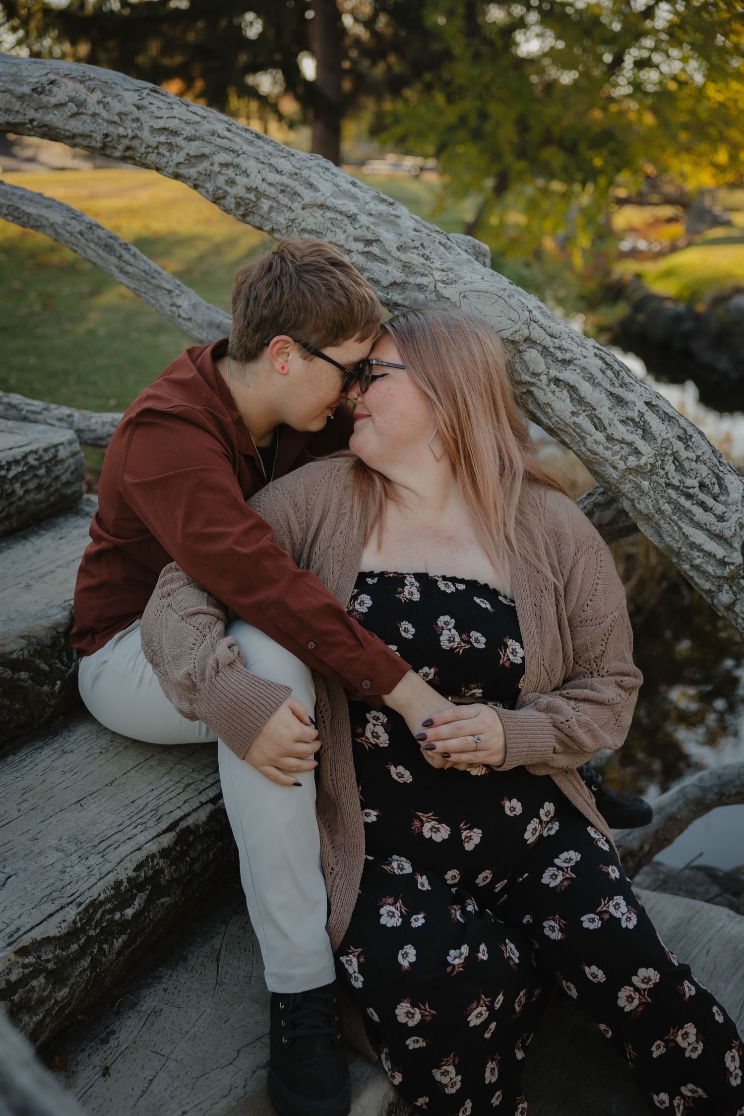 Couple sharing a romantic moment sitting on logs outdoors in autumn, woman in floral dress and man in red hoodie.