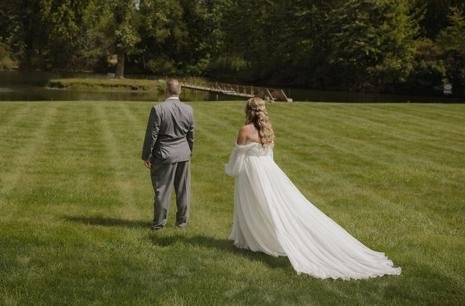 A couple in wedding attire walks hand in hand across a lush green lawn on a sunny summer day.