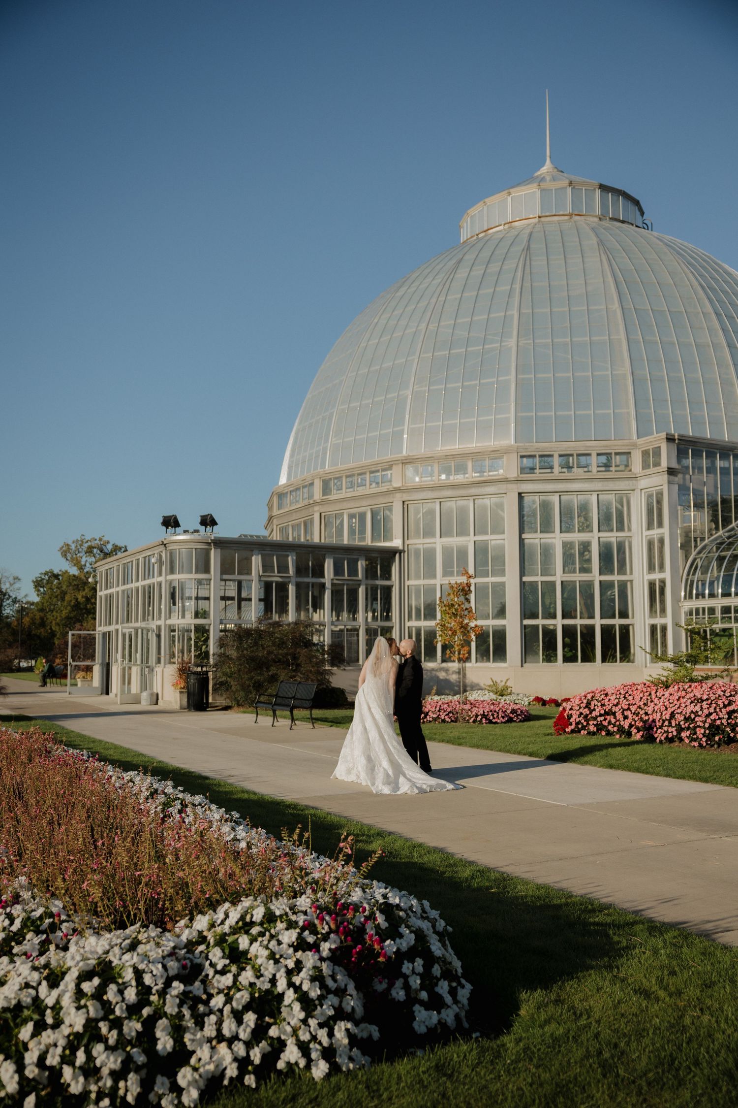 Bride and groom share a romantic moment outside a grand glass conservatory greenhouse surrounded by colorful flower gardens.
