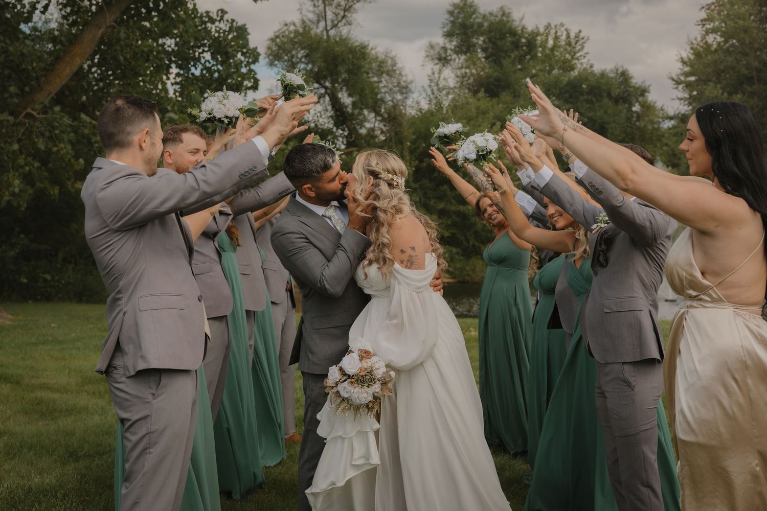 Wedding party in emerald green dresses and gray suits jumps joyfully on grass during outdoor celebration.
