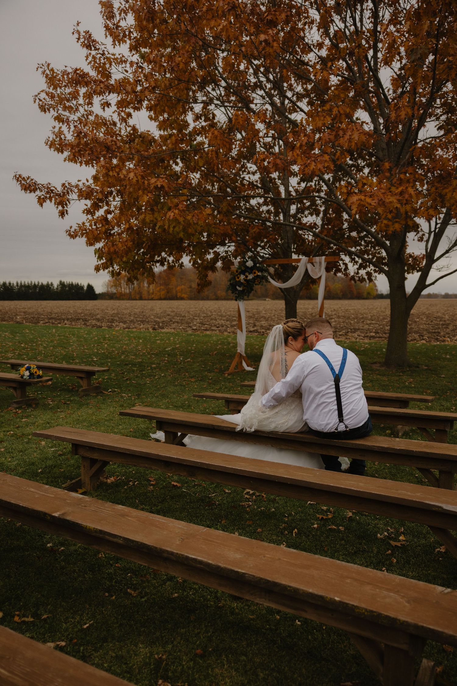 Bride and groom sit together on wooden benches at an outdoor fall wedding ceremony beneath an orange-leafed tree.