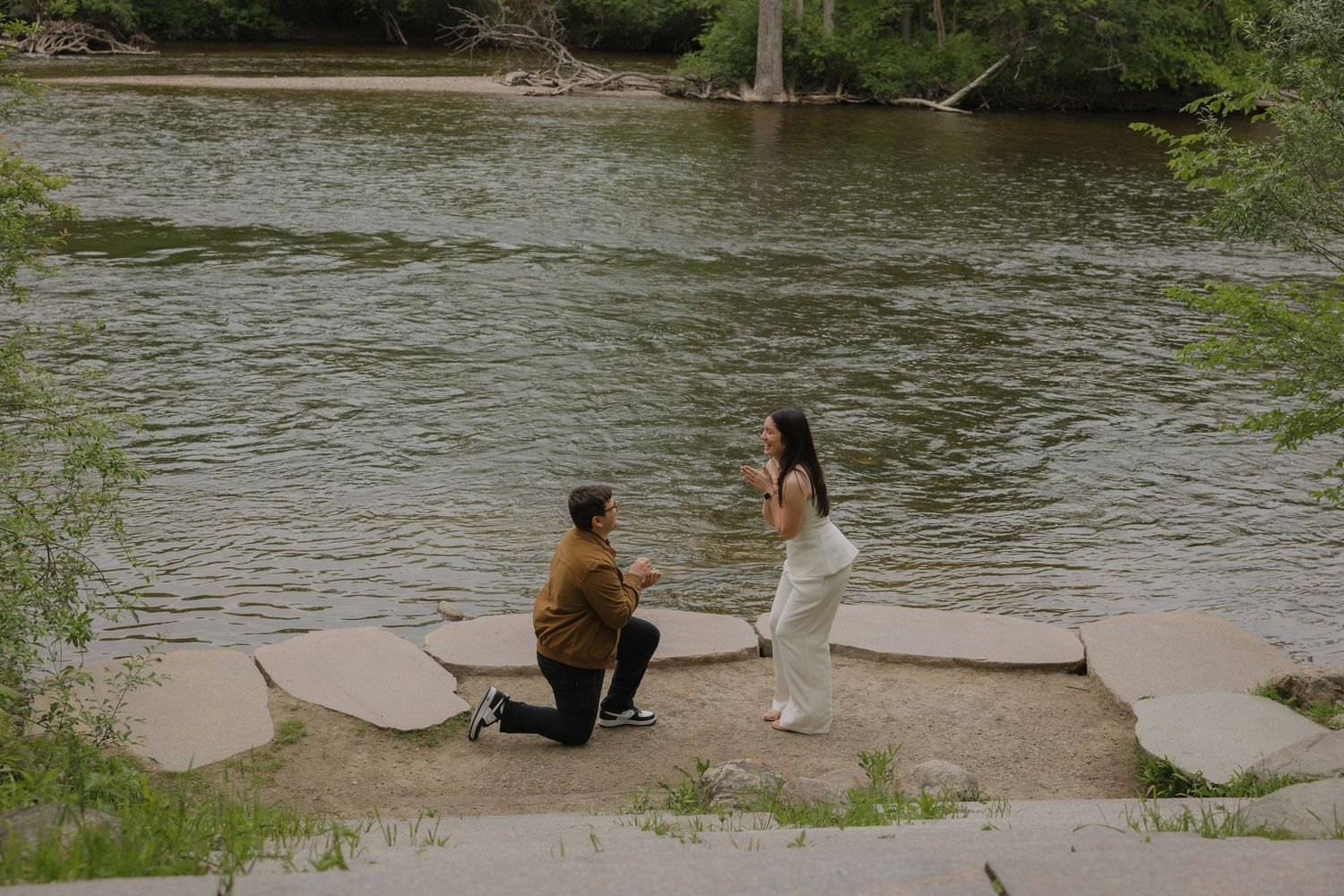 Man kneeling in marriage proposal to woman by a scenic river surrounded by lush green trees.