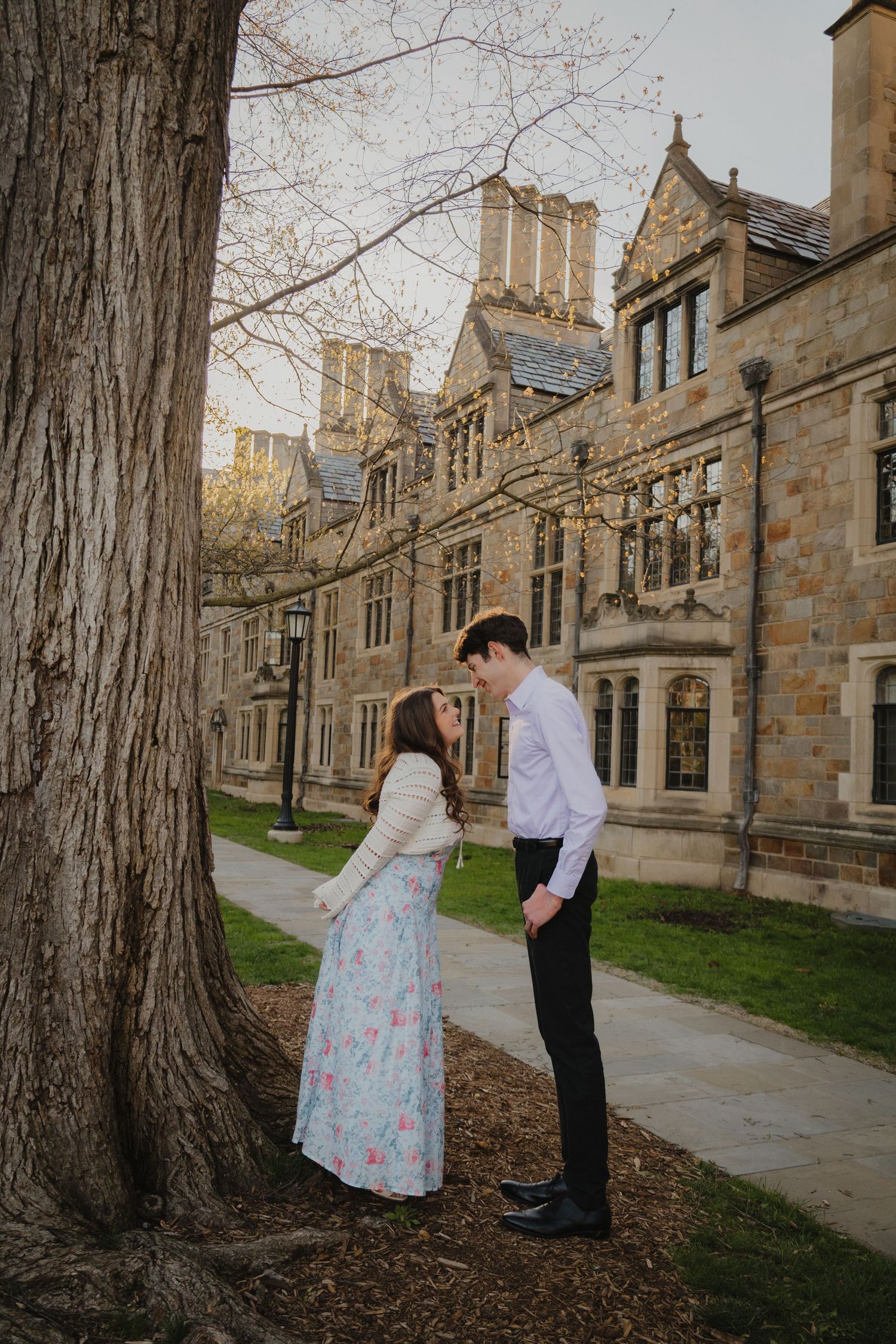 Couple sharing a romantic moment by a tree outside a historic stone university building in spring.