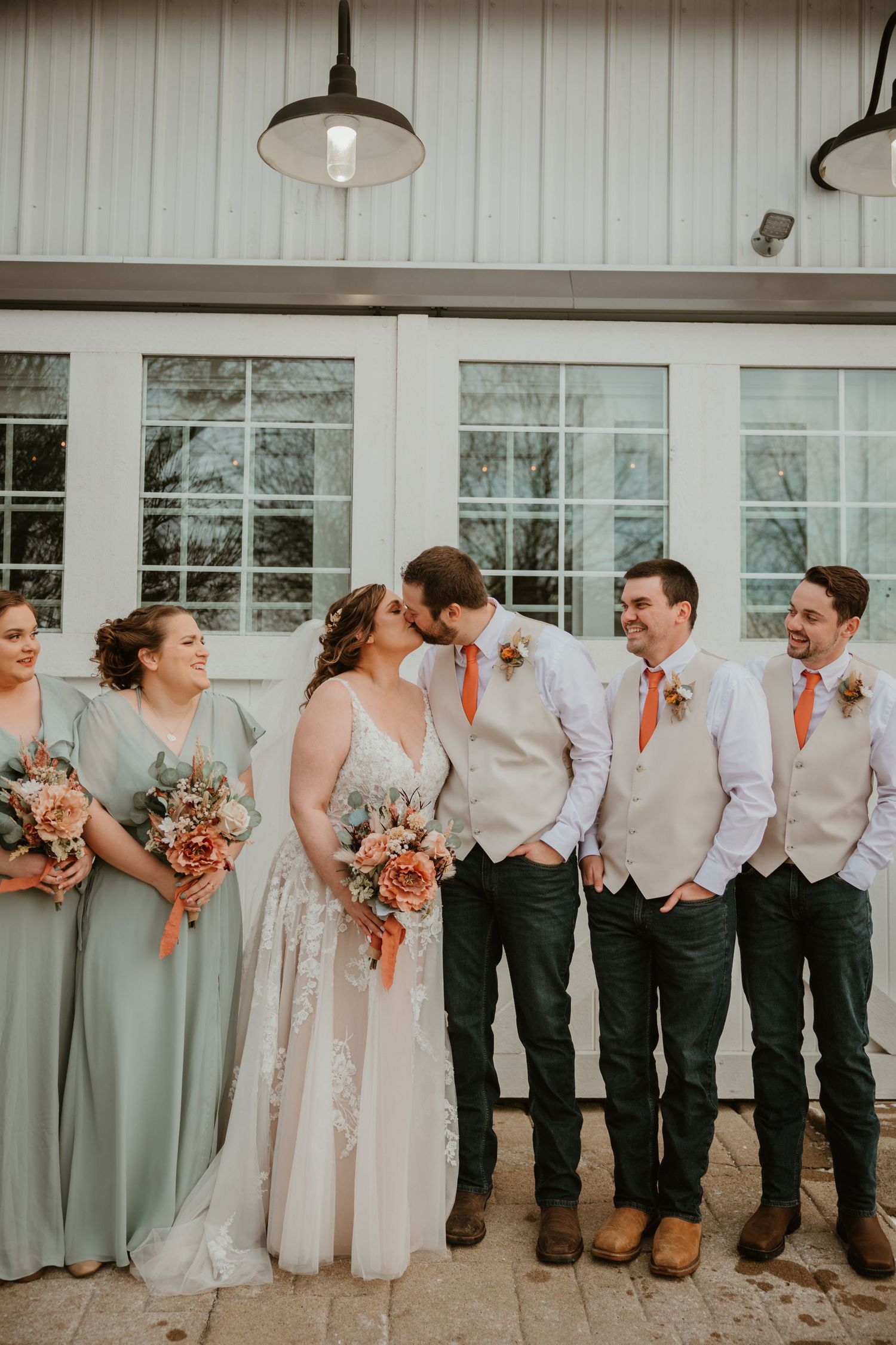 Wedding party in sage green dresses and light gray vests pose together against white building with windows.