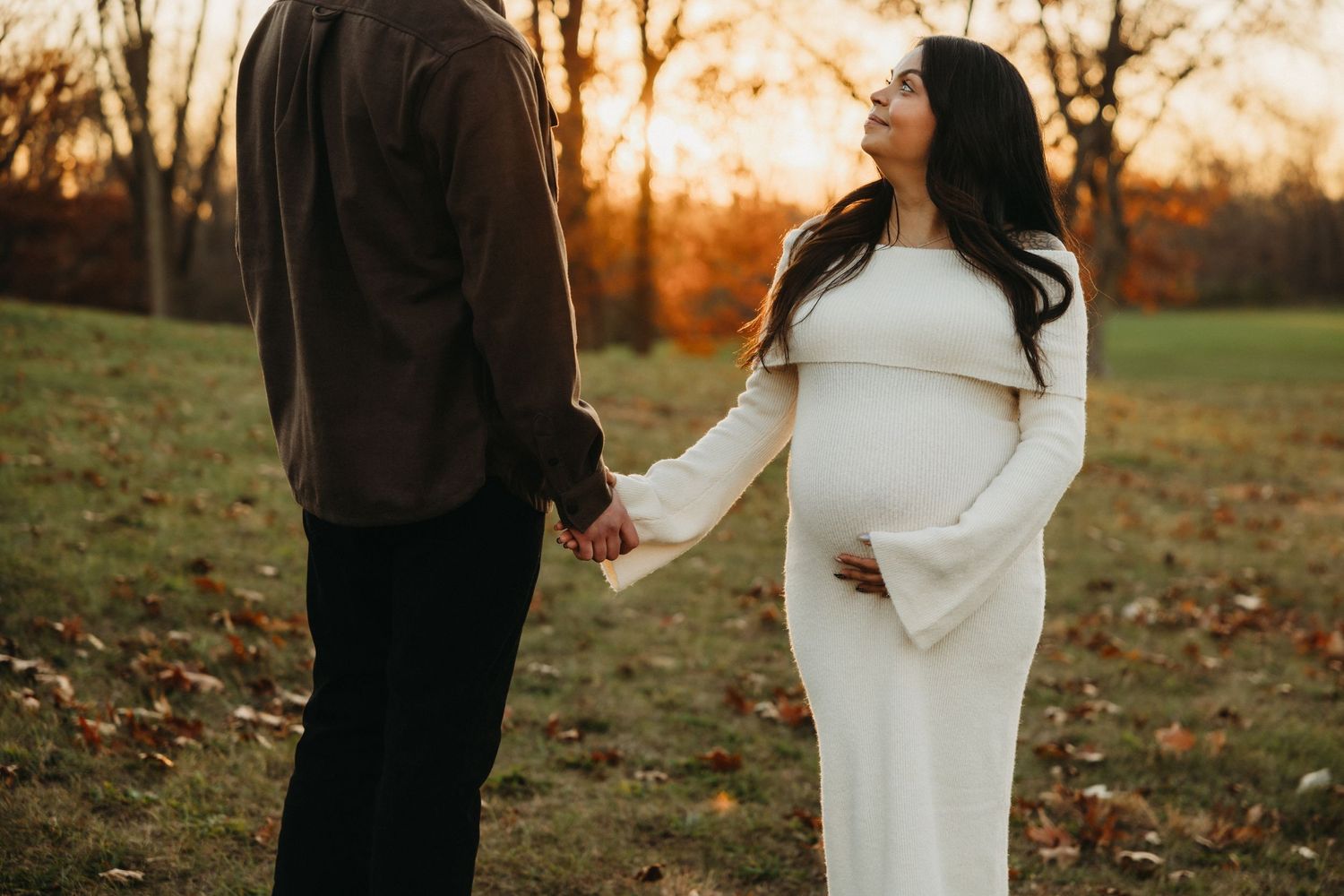 Couple stands in autumn field during maternity photo shoot at sunset, with woman wearing white dress.