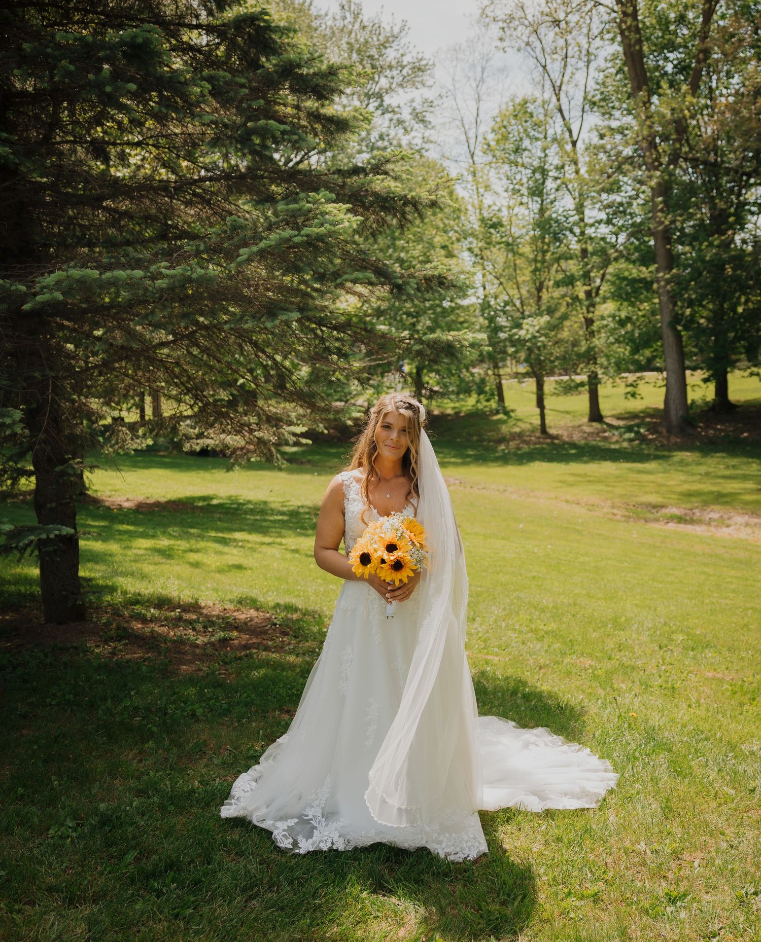 Bride in white lace gown and veil holding sunflower bouquet standing on green lawn surrounded by trees.