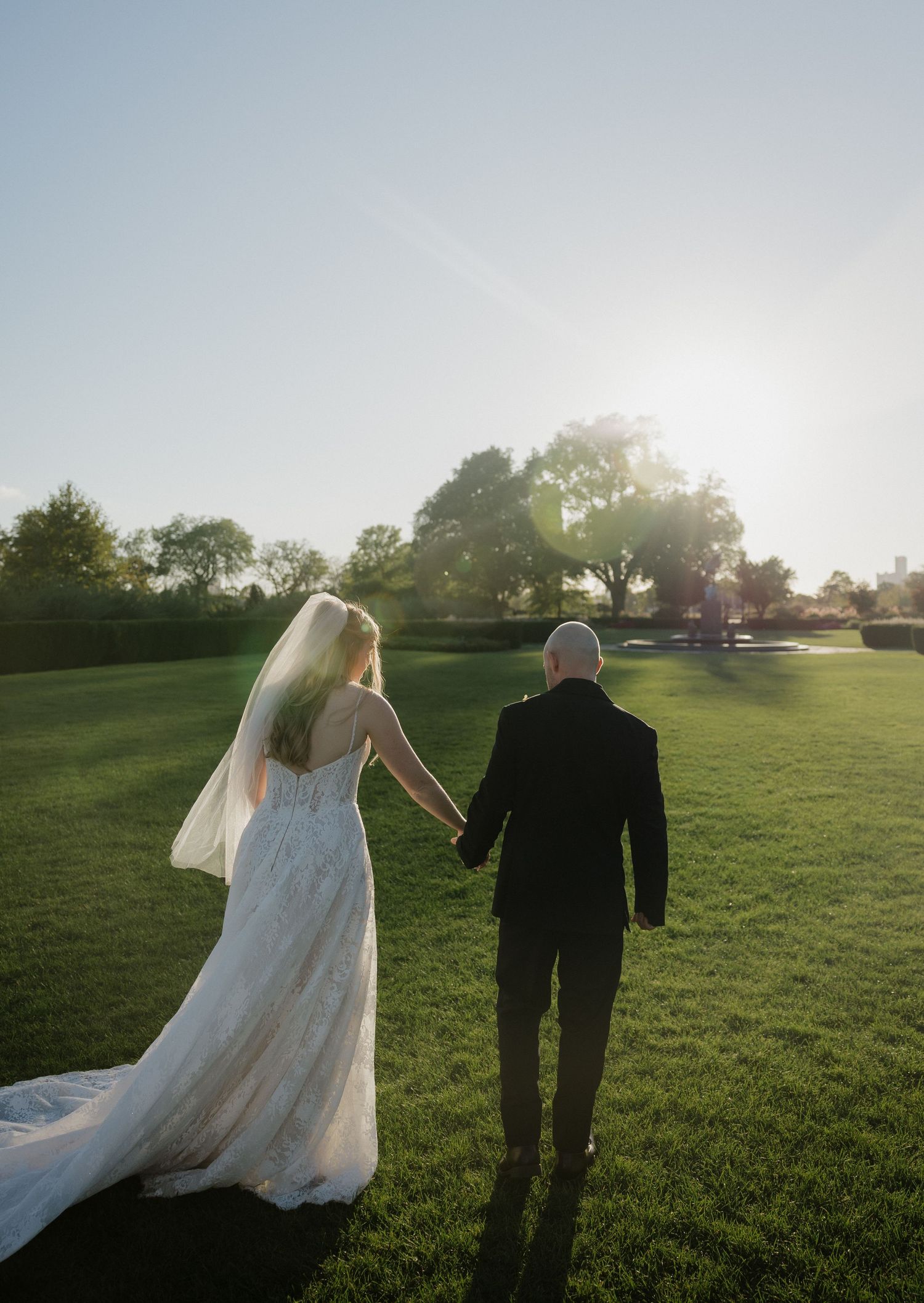 Bride and groom walking hand in hand across lush green lawn at golden hour sunset wedding celebration.