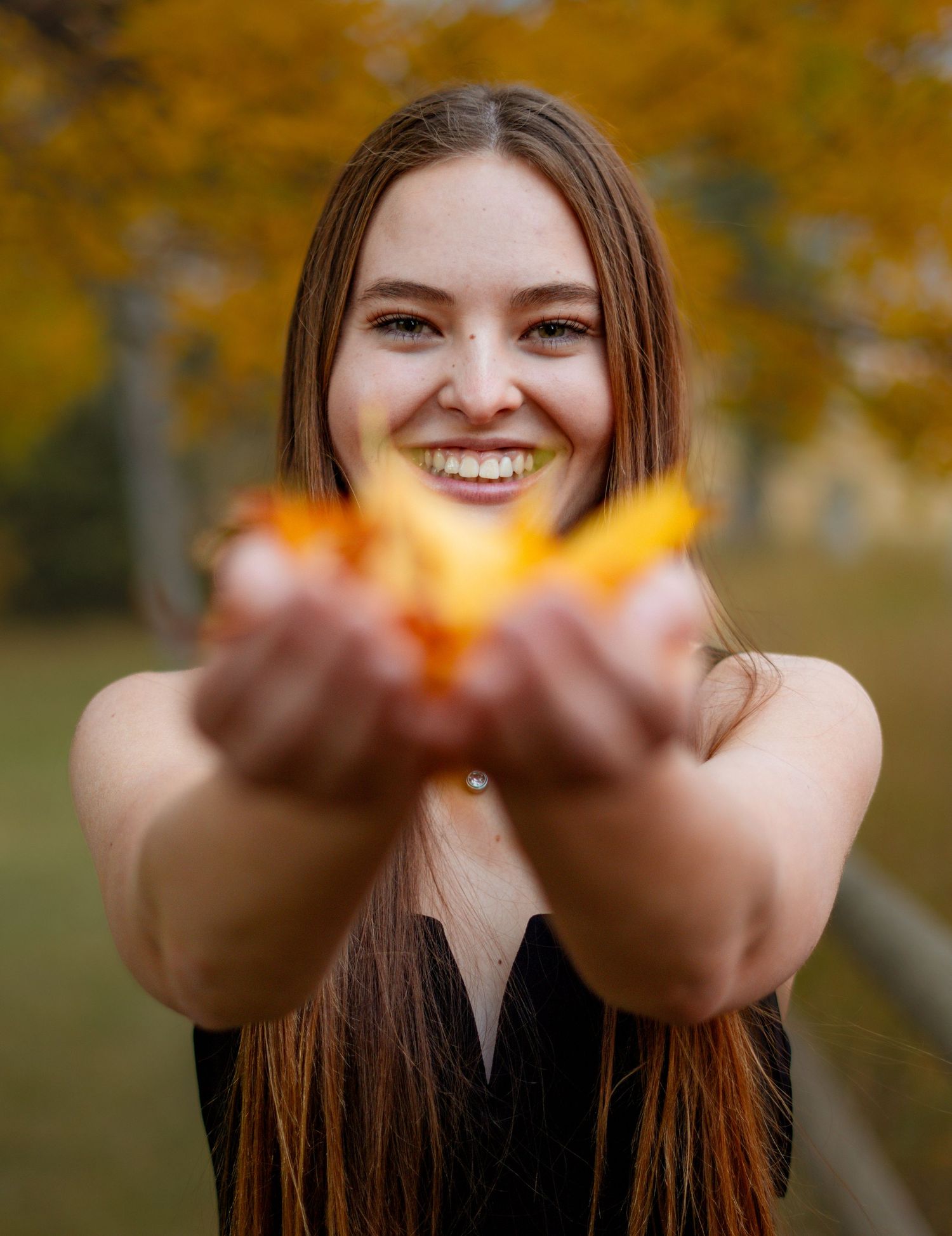 A smiling person with long brown hair holds autumn leaves in outstretched hands against a blurred fall background.
