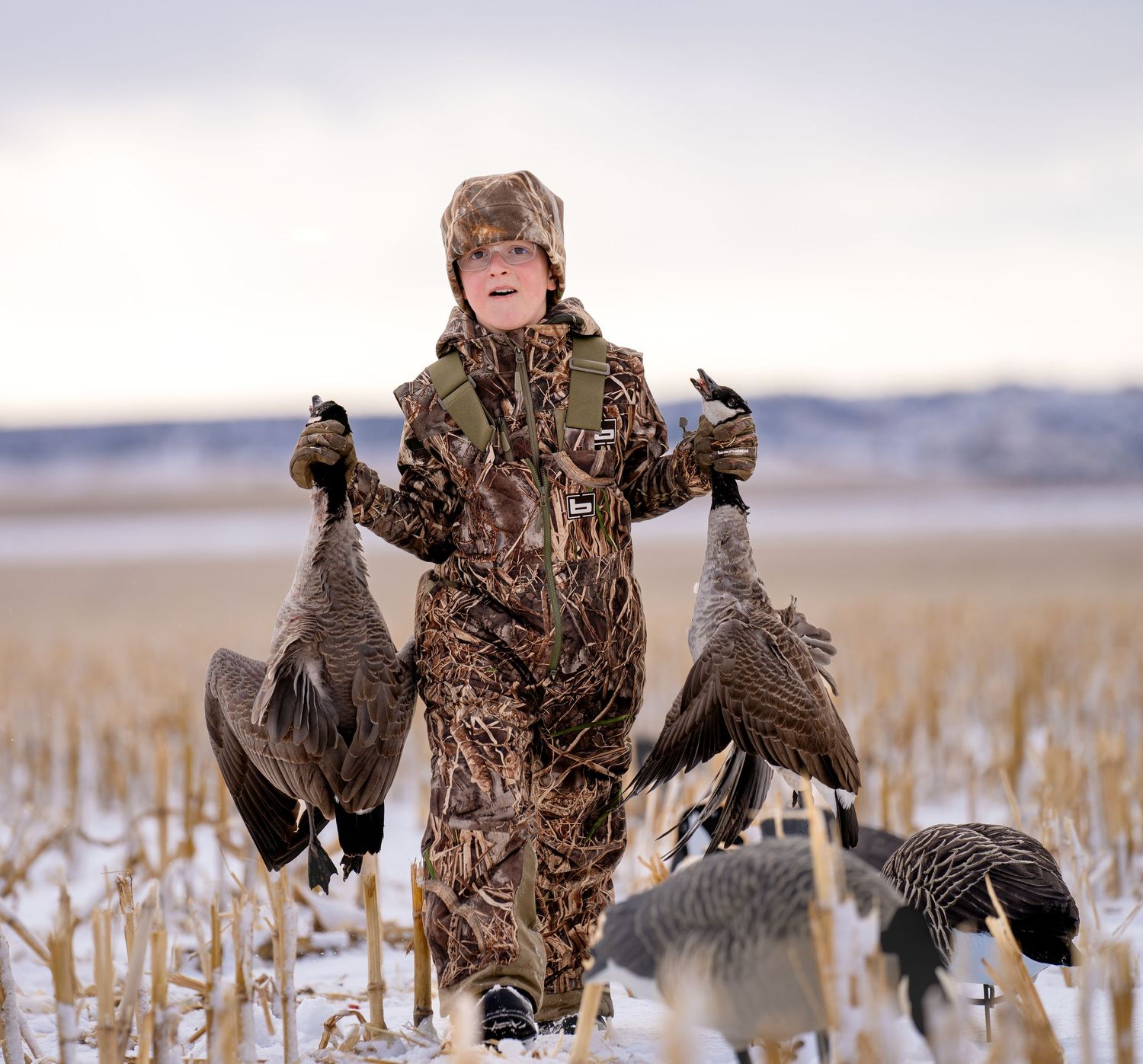 Hunter with camouflage clothing holds up two harvested geese in a snowy field while retrieving game birds.