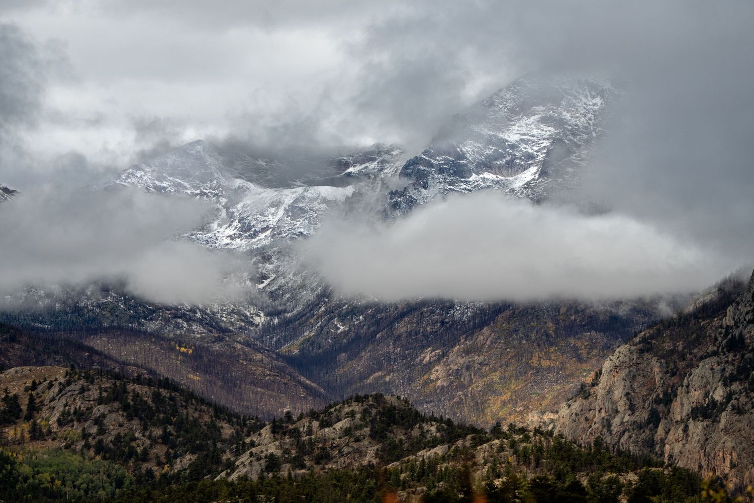 Snow-capped mountain peaks emerge through dramatic storm clouds over rugged rocky terrain in autumn.