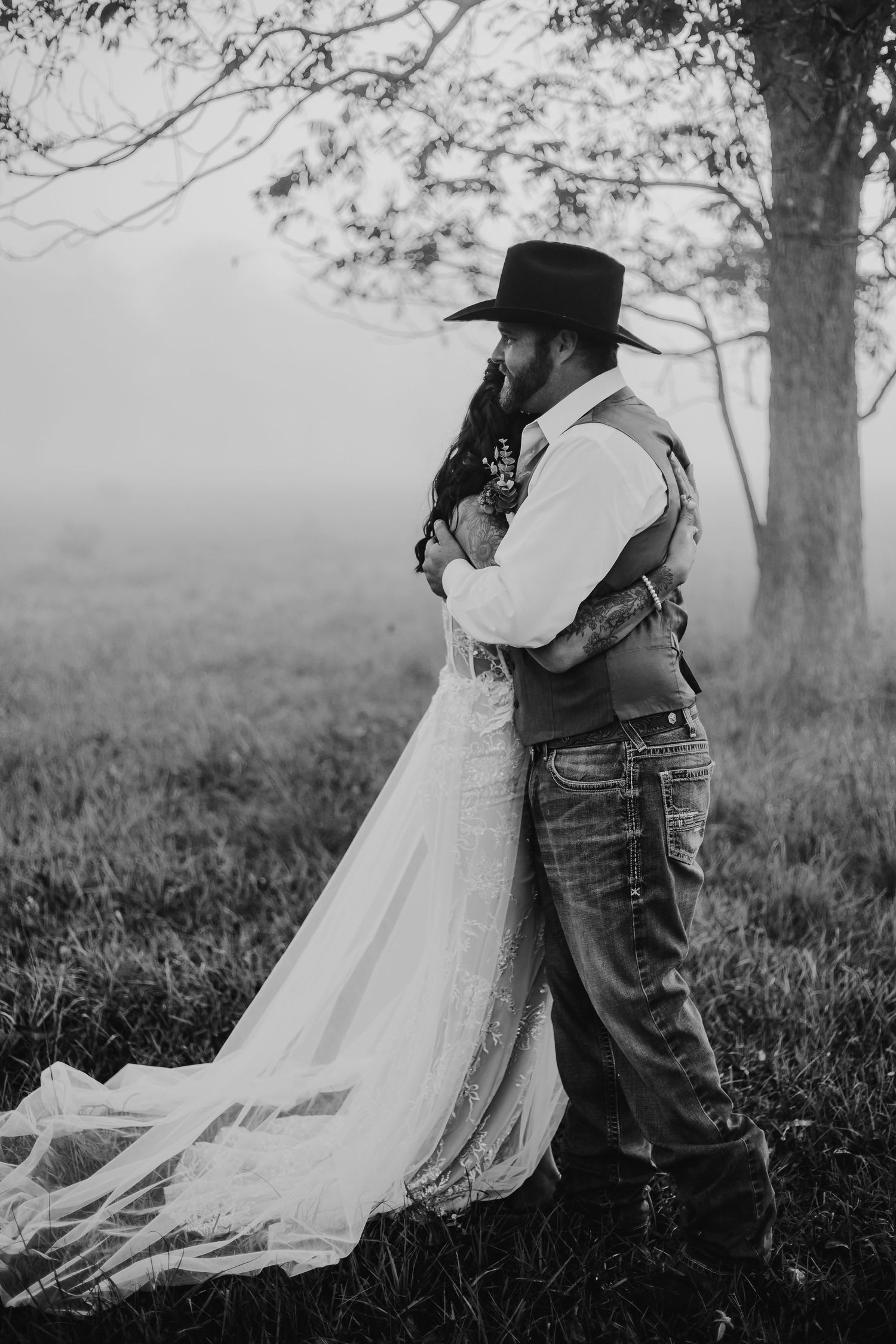 A romantic black and white portrait of a couple embracing under a tree in a misty field.