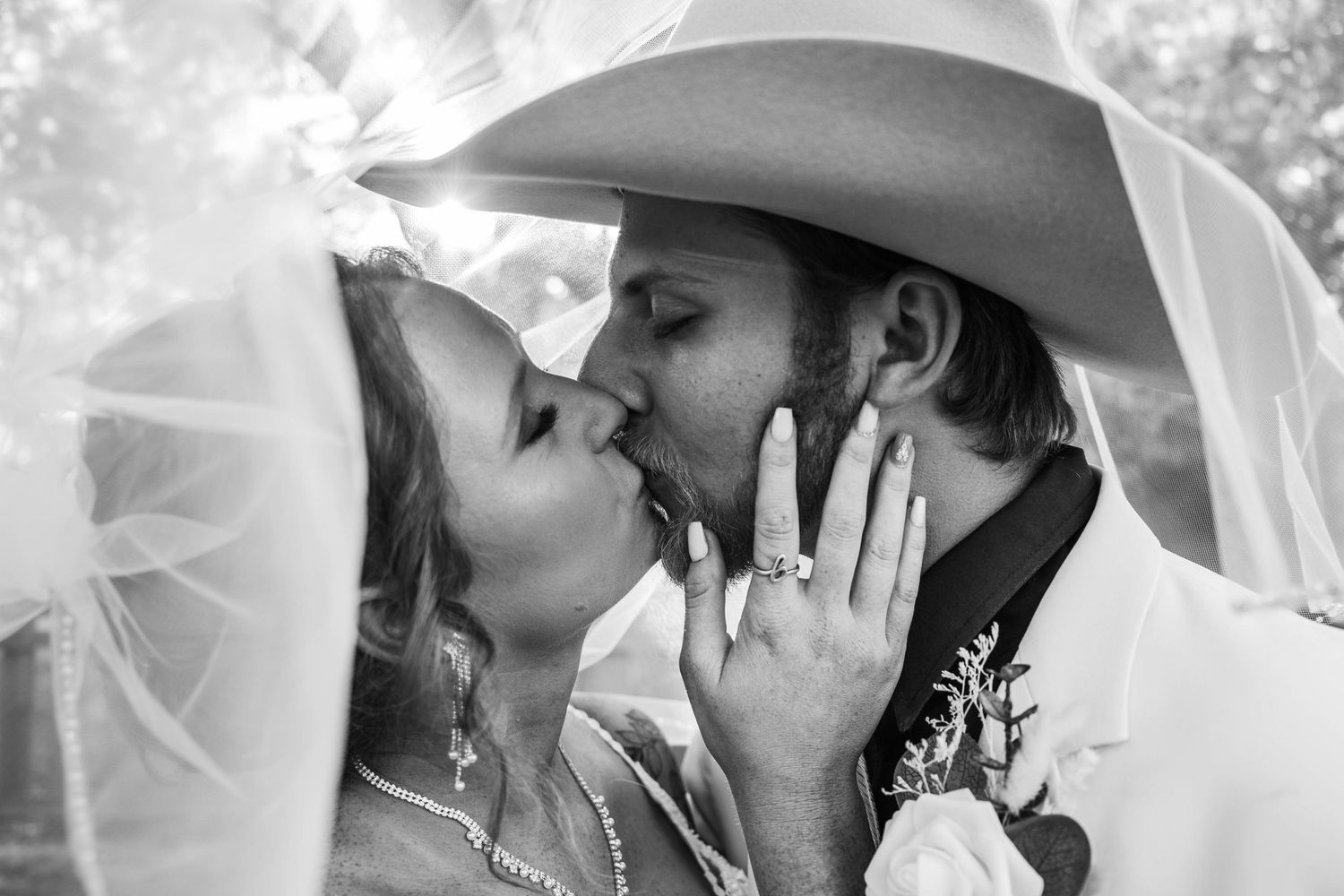 A black and white close-up of newlyweds sharing a kiss under a cowboy hat and bridal veil.