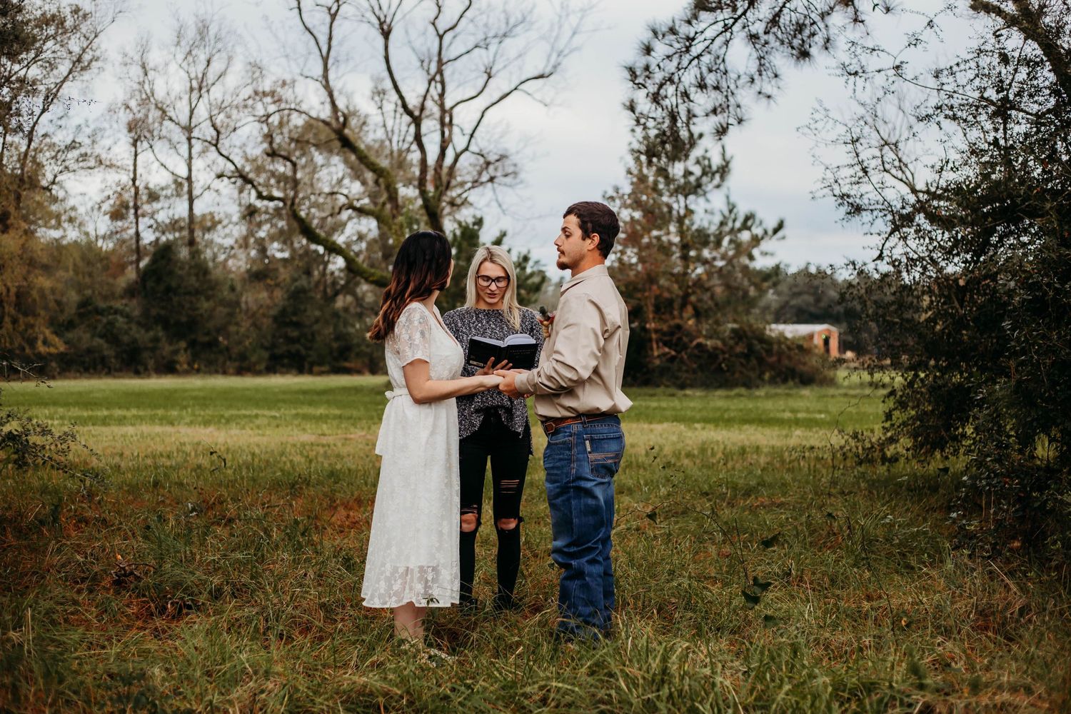 A rustic outdoor wedding ceremony takes place in a field with bare trees in the background.