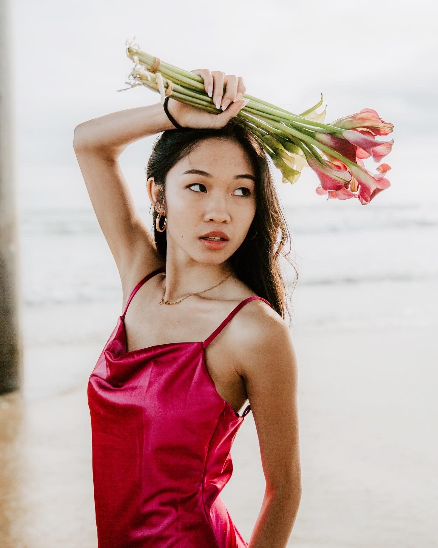 Woman in red dress poses gracefully with flowers on a beach at sunset.