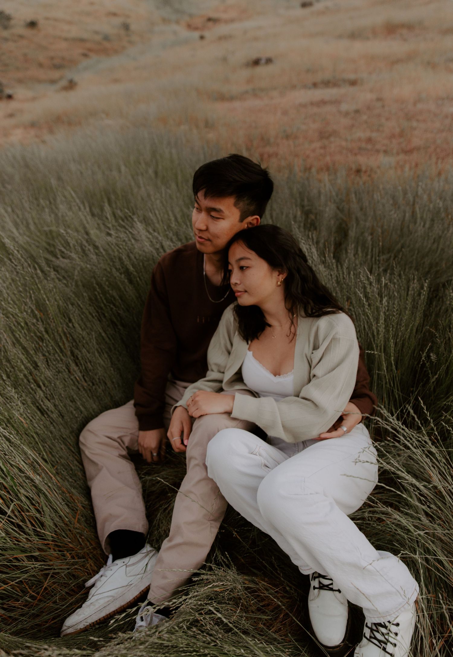 Couple in casual outfits sitting together in tall grass during sunset.
