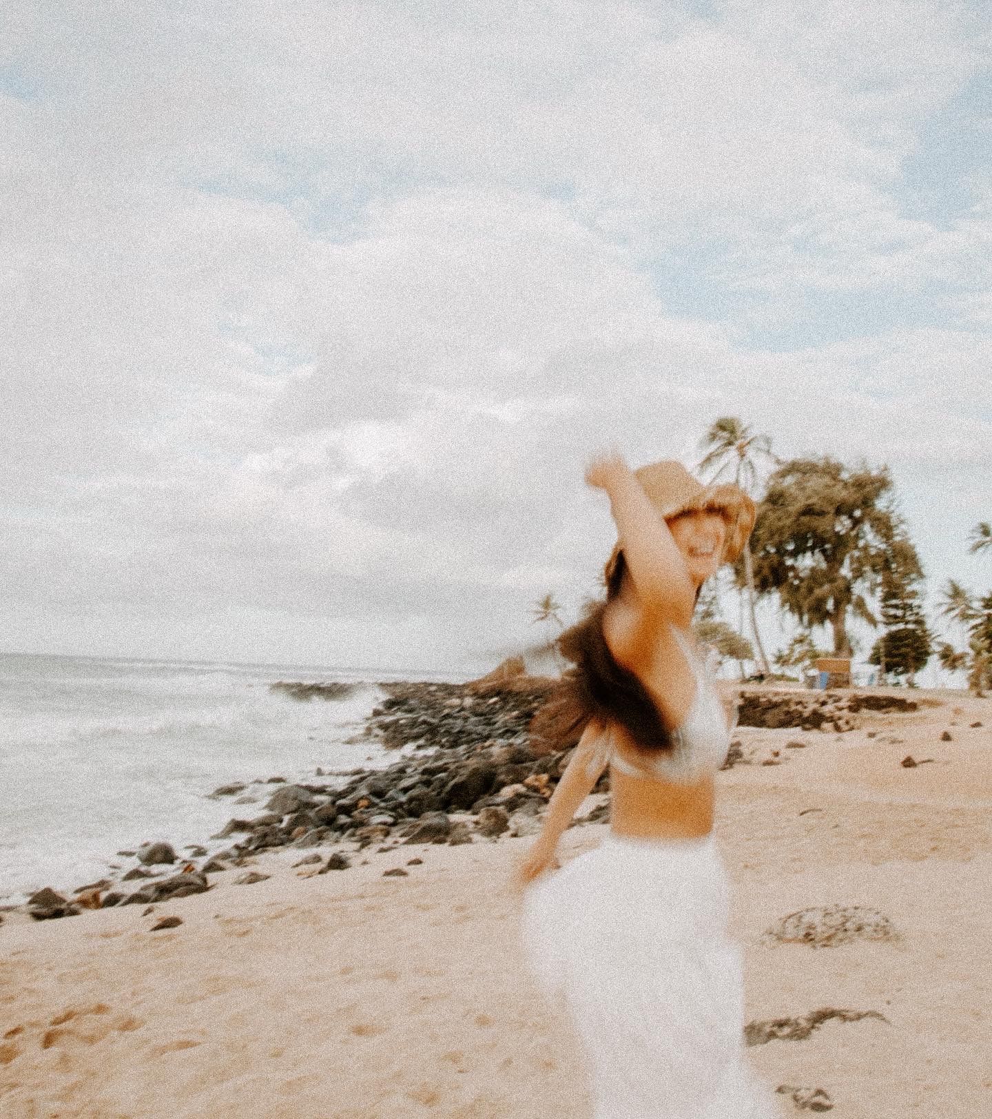 Person in white dress twirling joyfully on a rocky beach with palm trees in the background.