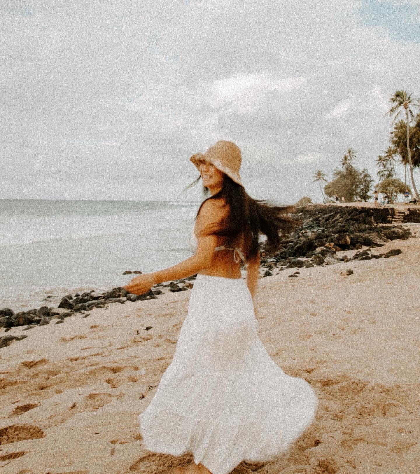 Person in white dress and sun hat twirling on a beach with palm trees at sunset.