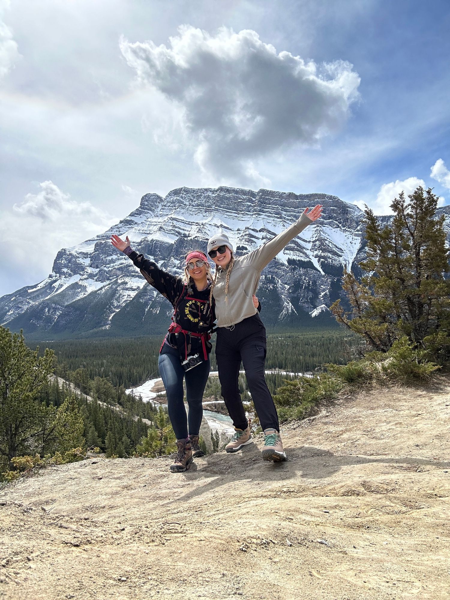 Two excited people jump with outstretched arms on a mountain viewpoint with snow-capped Mount Rundle in Banff behind them.