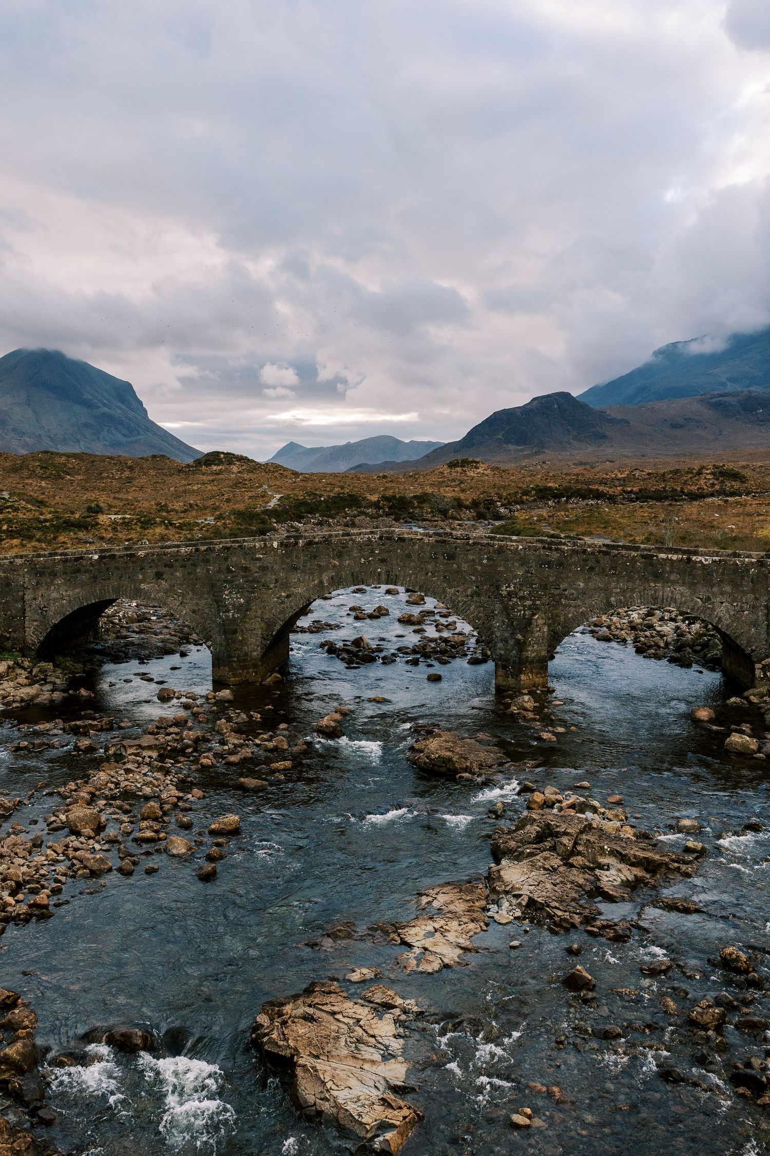 Ancient stone bridge spans rocky stream with misty Scottish Highlands mountains in background at Sligachan, Isle of Skye.