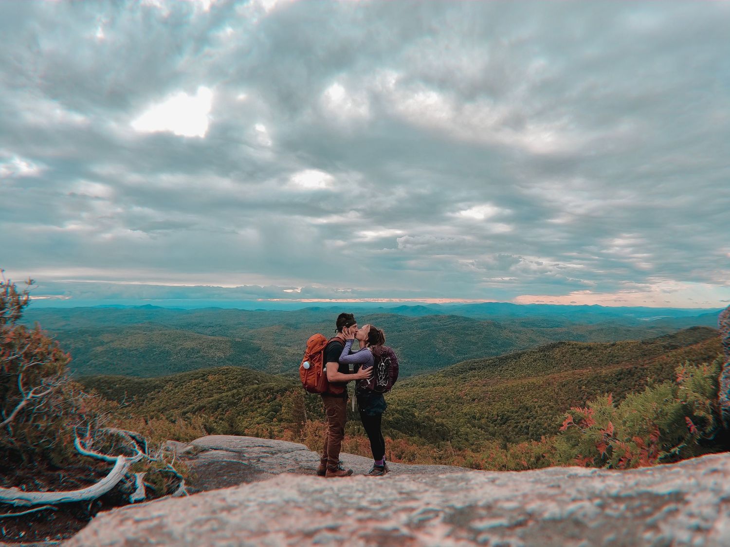 Two hikers enjoy a scenic mountain vista with dramatic clouds and rolling hills stretching to the horizon.