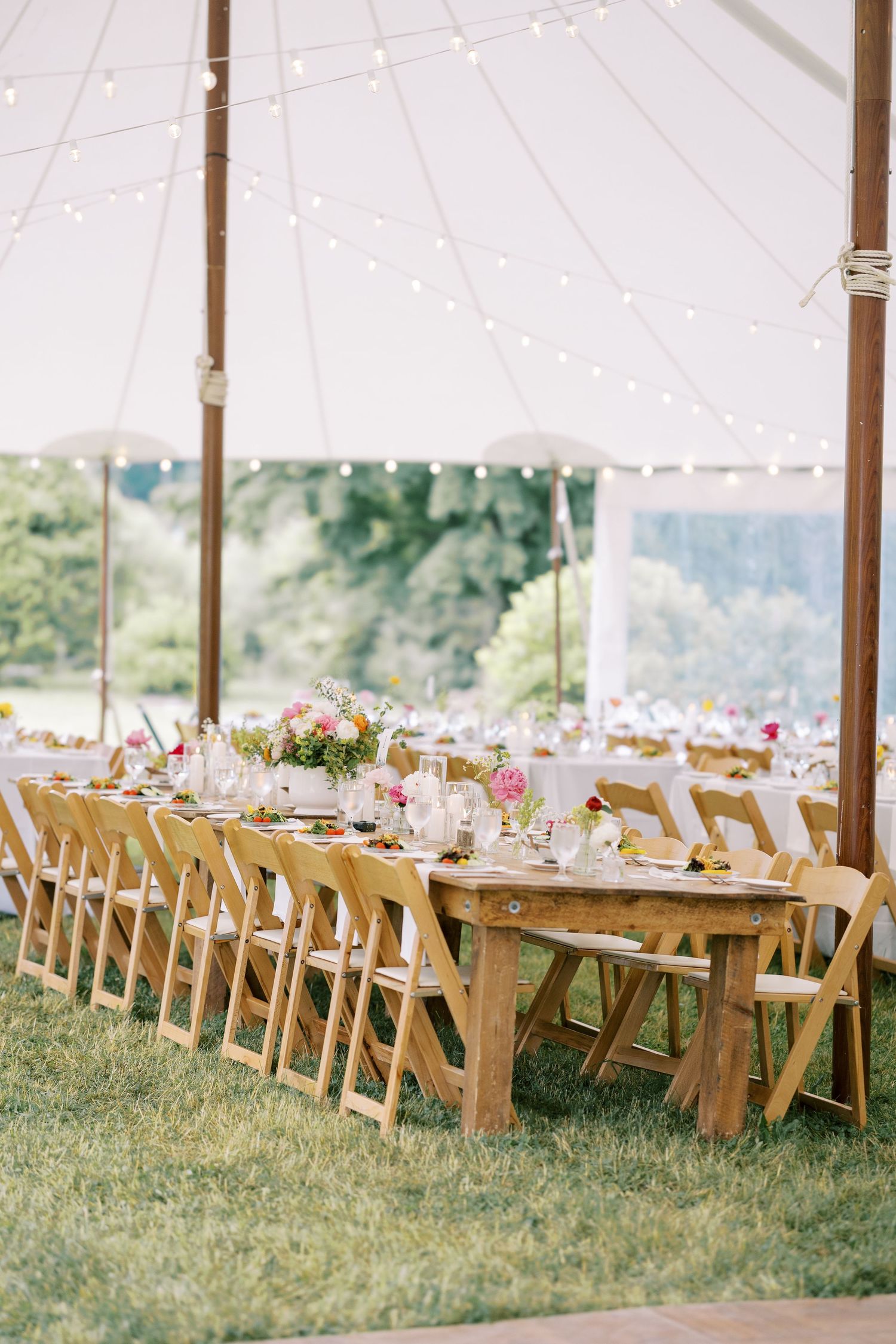 Long rows of wooden chairs and tables set for a rustic outdoor wedding reception under a tent on grassy lawn with floral decor.