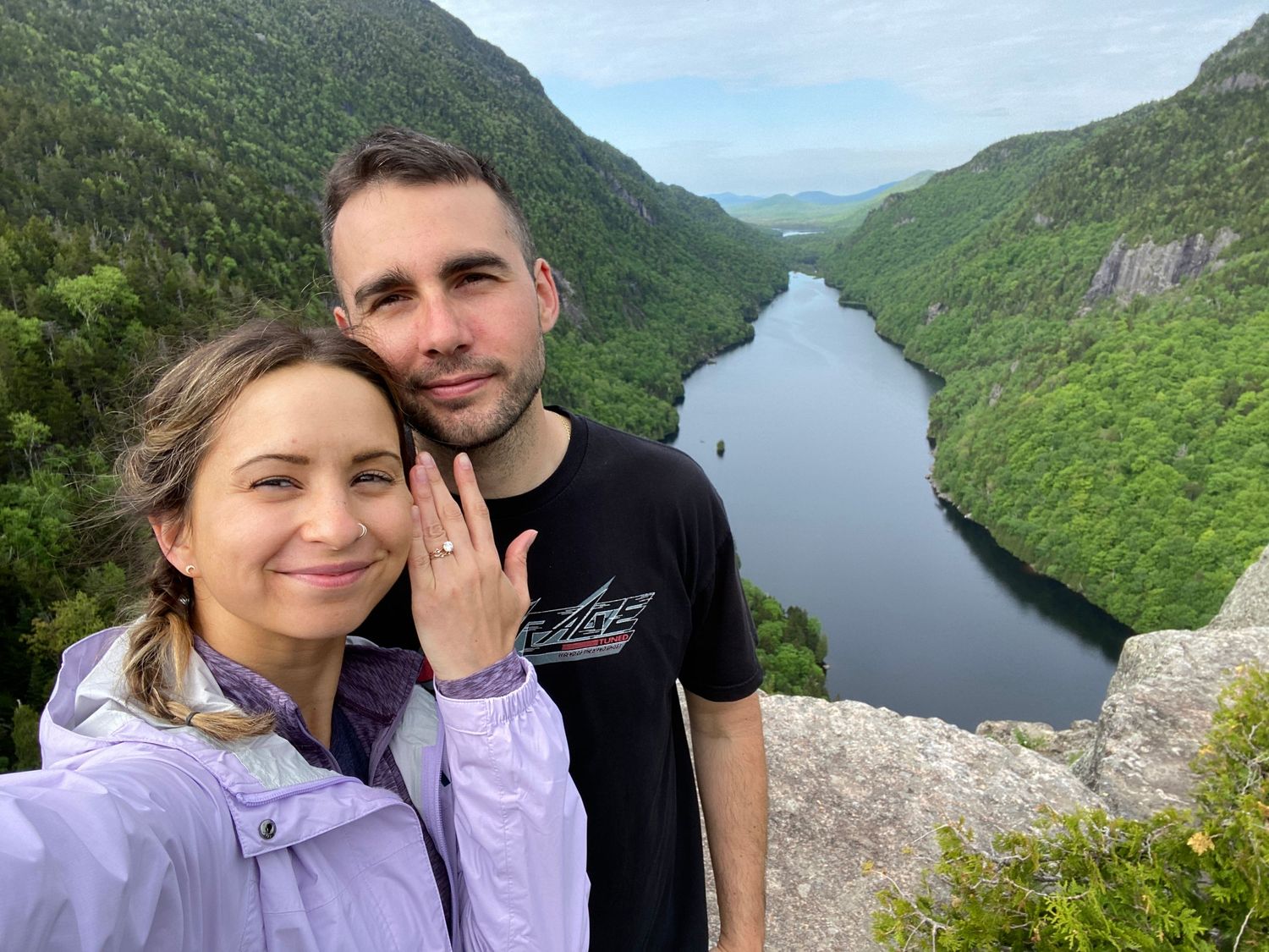 Couple stands at scenic mountain viewpoint overlooking winding river valley through mountainous wilderness landscape.
