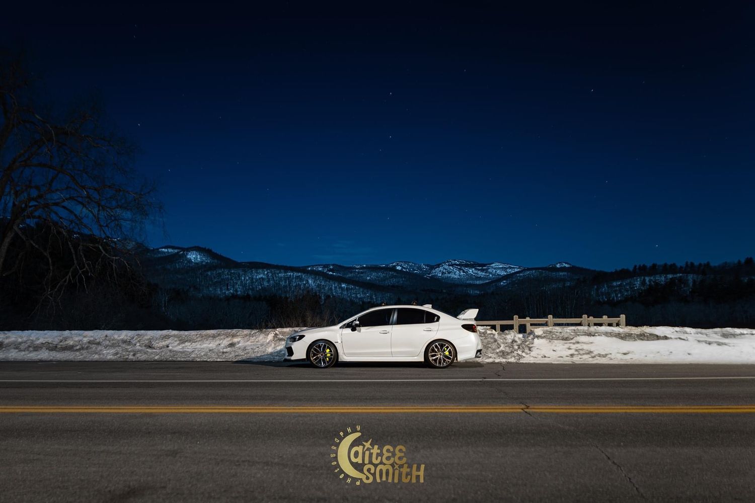 White sports car parked on snowy mountain road against blue sky and snow-capped peaks in winter landscape.