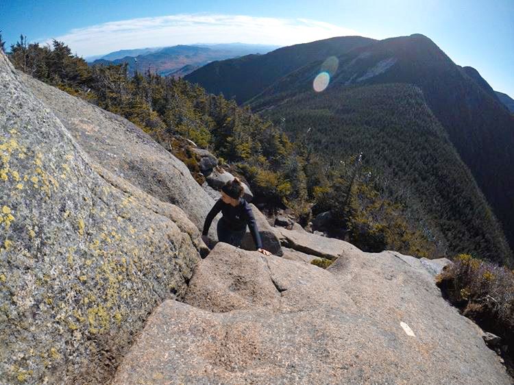 A rock climber rests on a steep granite slope with expansive mountain views and forested valleys below.