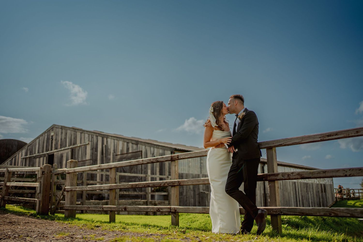 Rustic barn wedding portrait with couple embracing against wooden fence and blue sky.