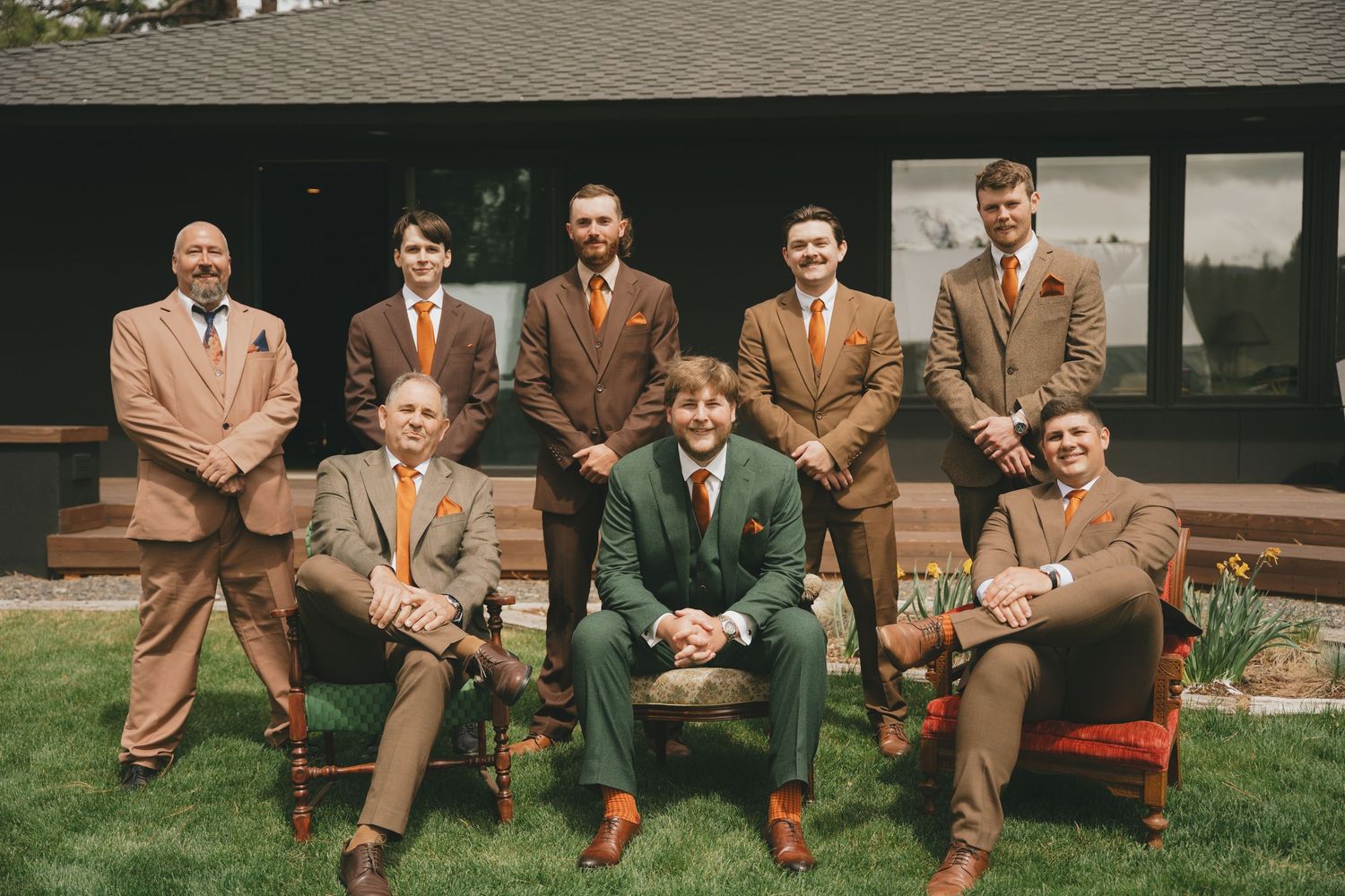 Groomsmen in tan and brown suits pose on a wooden bench outdoors for wedding party photos.