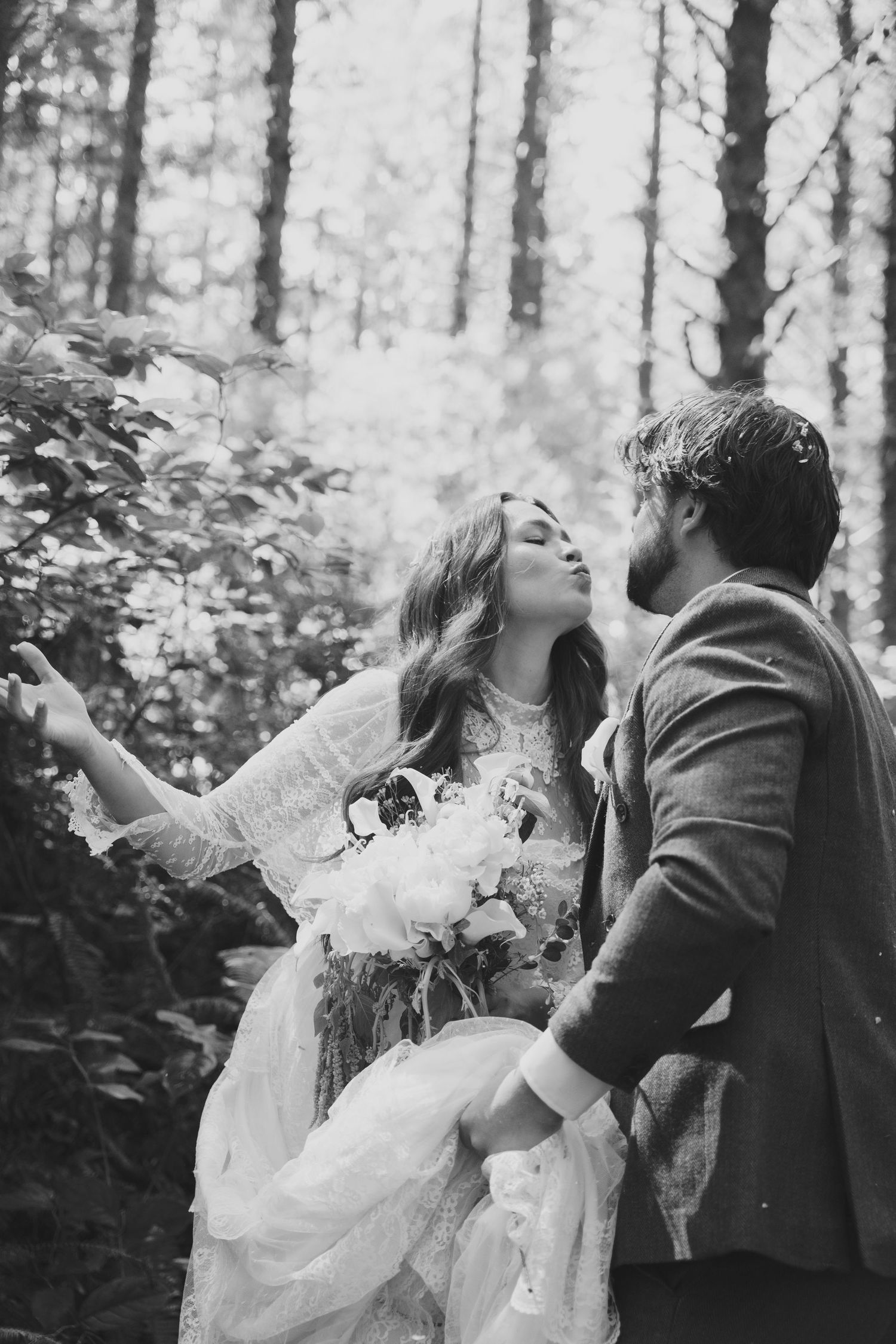 Black and white photo of a bride and groom sharing a romantic moment in a lush forest, bride holding a bouquet.
